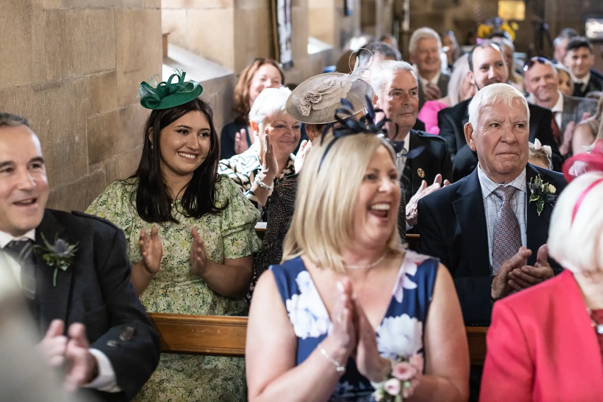 A group of well-dressed people sit on pews, clapping and smiling, at what appears to be a formal event inside a building with stone walls.