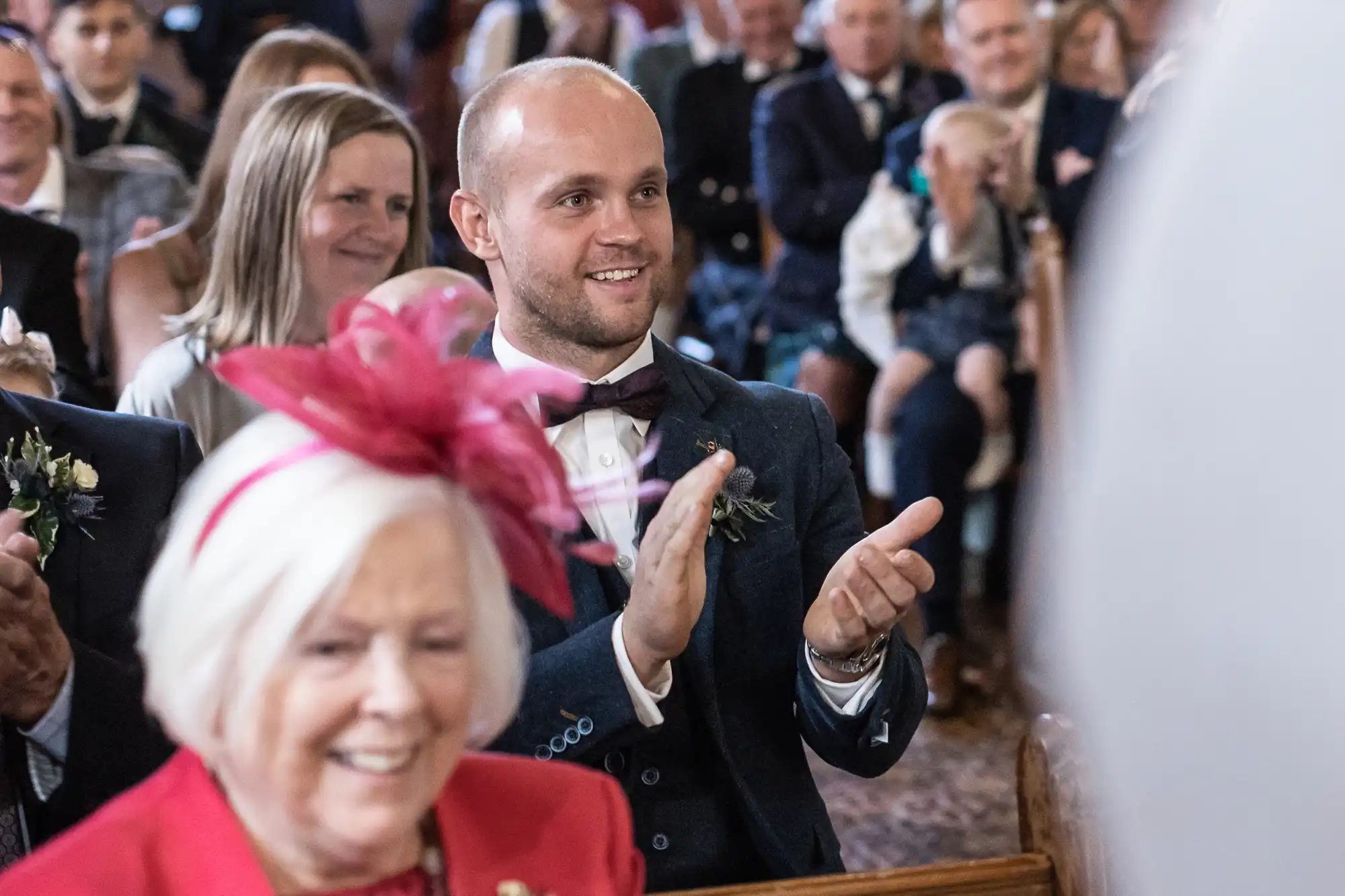 A man in formal attire claps while seated among other people at a gathering, with an older woman wearing a red outfit and pink hat in the foreground.