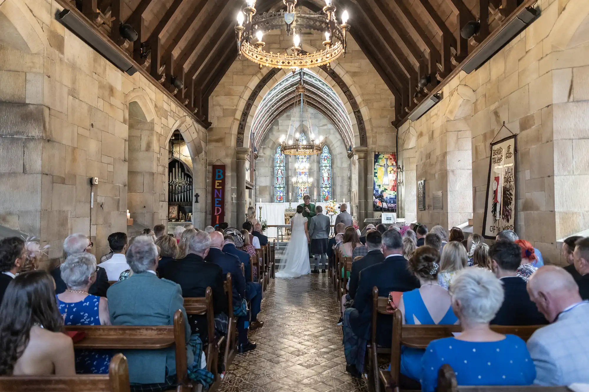 A couple stands at the altar during a church wedding ceremony, viewed from the back of the congregation seated in wooden pews in a stone chapel with a vaulted ceiling and stained glass windows.