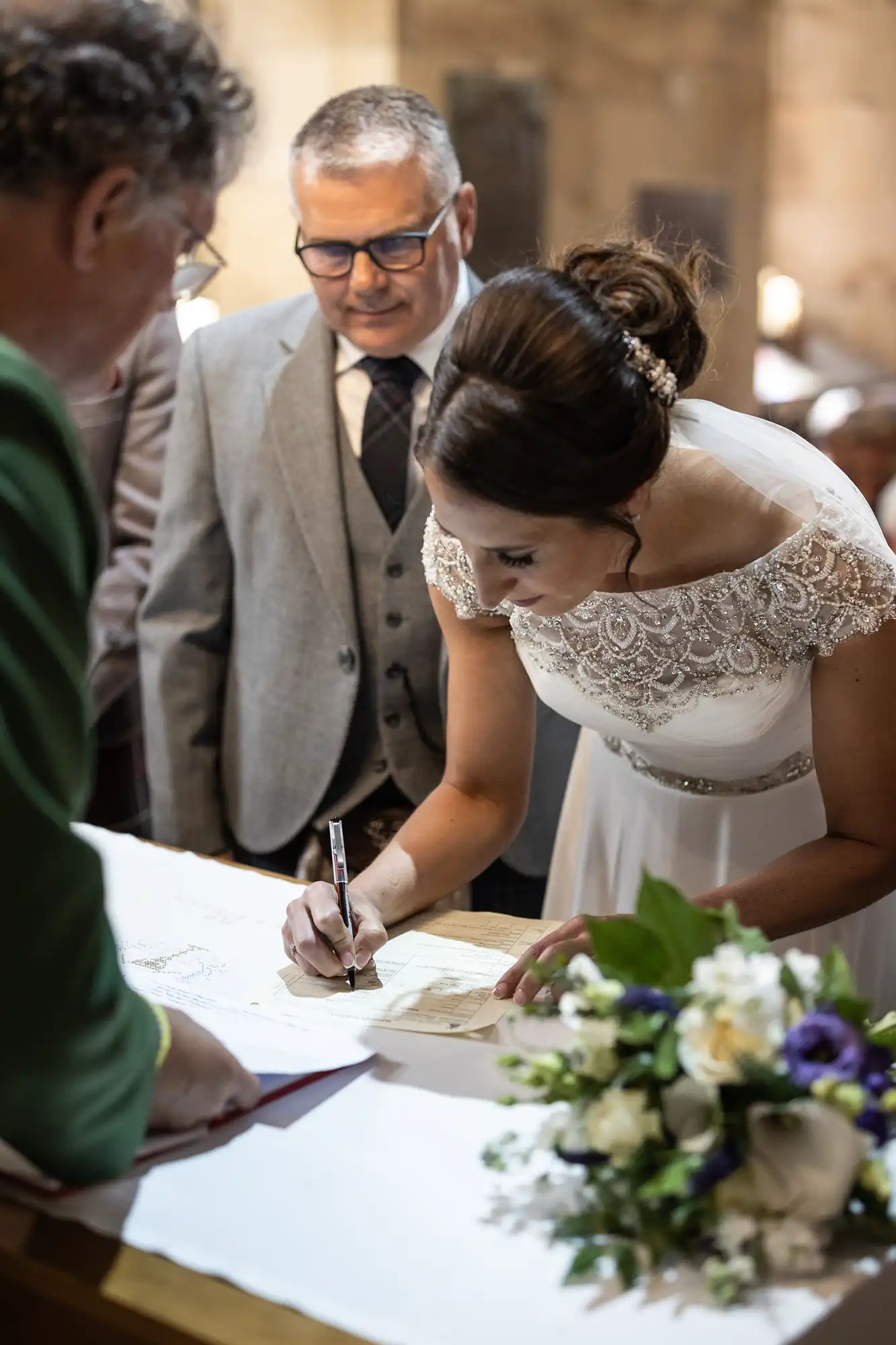 A bride in a white dress signs a document on a table while a groom in a suit and an officiant look on. Flowers are visible on the table.