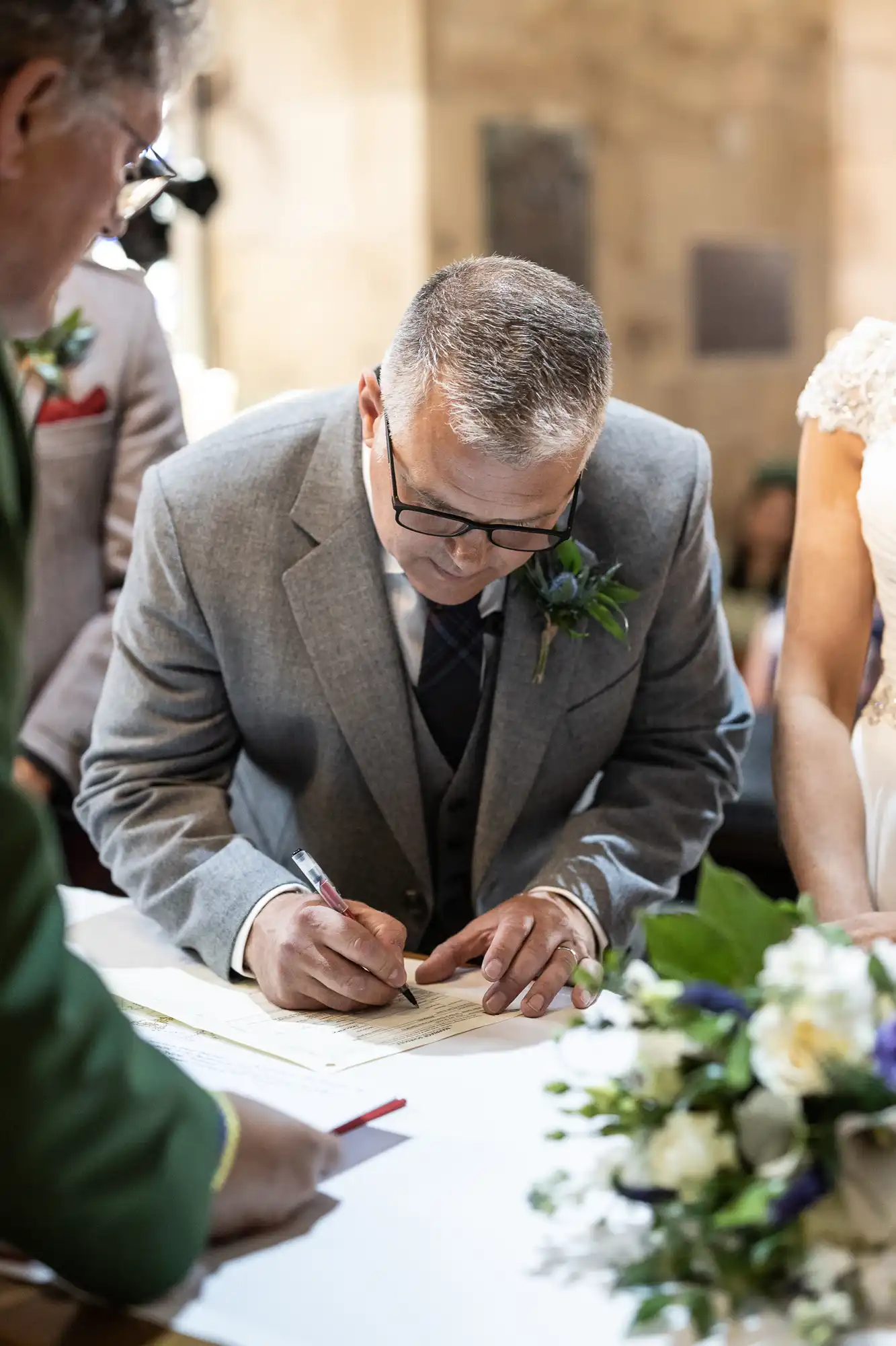 A man in a gray suit signs a document at a table with flowers, while a woman in a white dress stands beside him.