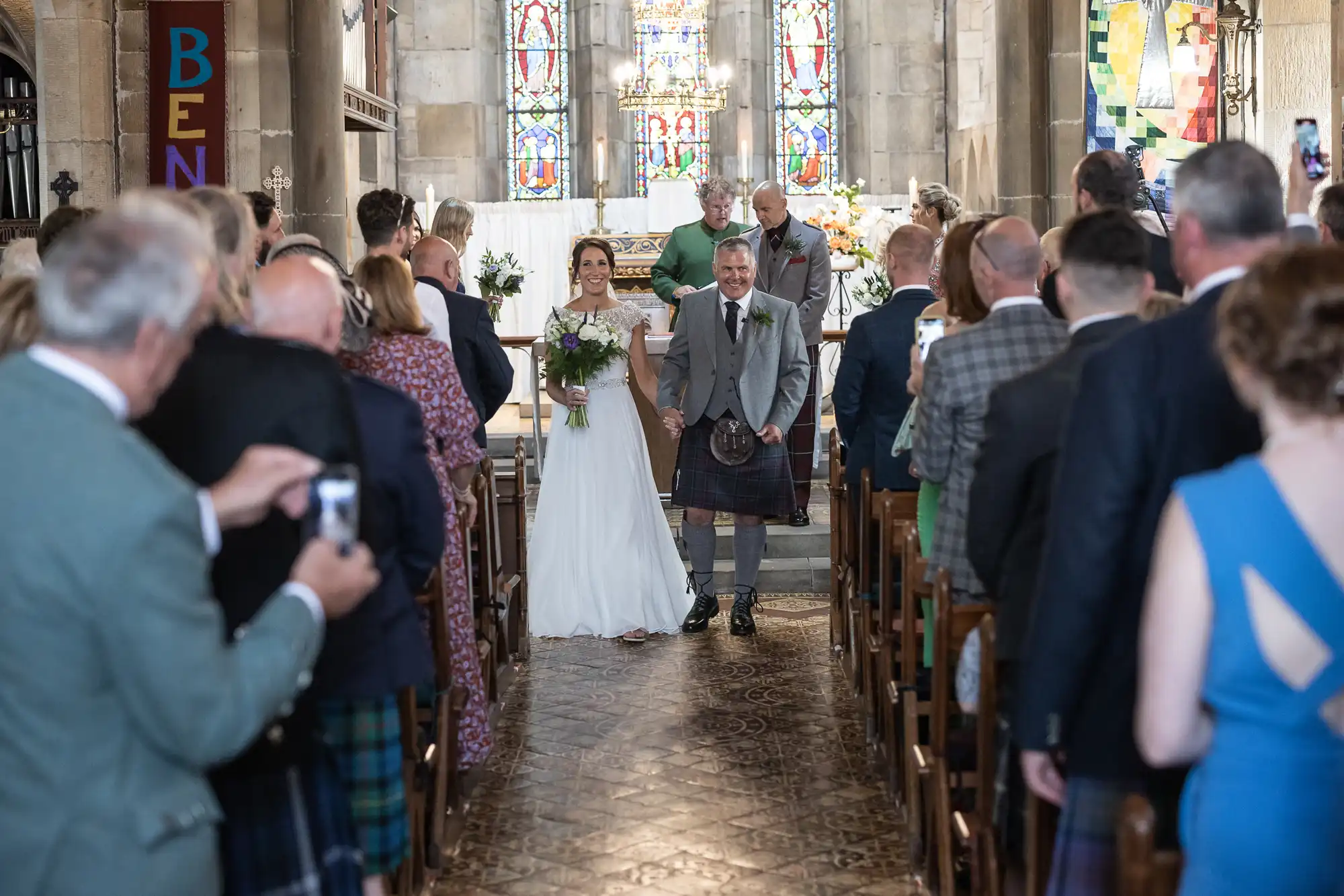 A bride walks down the church aisle escorted by an older man, with wedding guests standing on either side taking photos and watching. Stained glass windows and an altar are visible at the front.