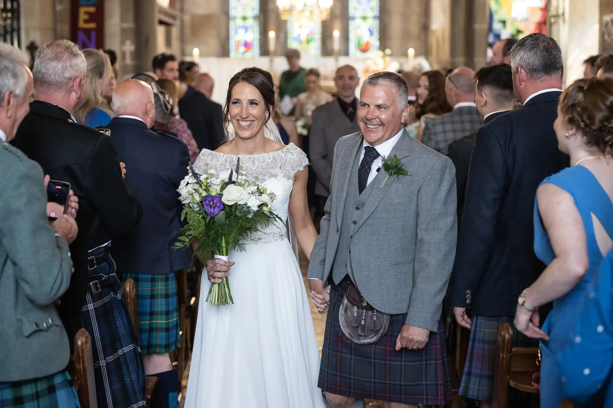 A bride and groom holding hands while walking down the aisle in a church, surrounded by guests. The groom wears a kilt, and the bride holds a bouquet of white and purple flowers.