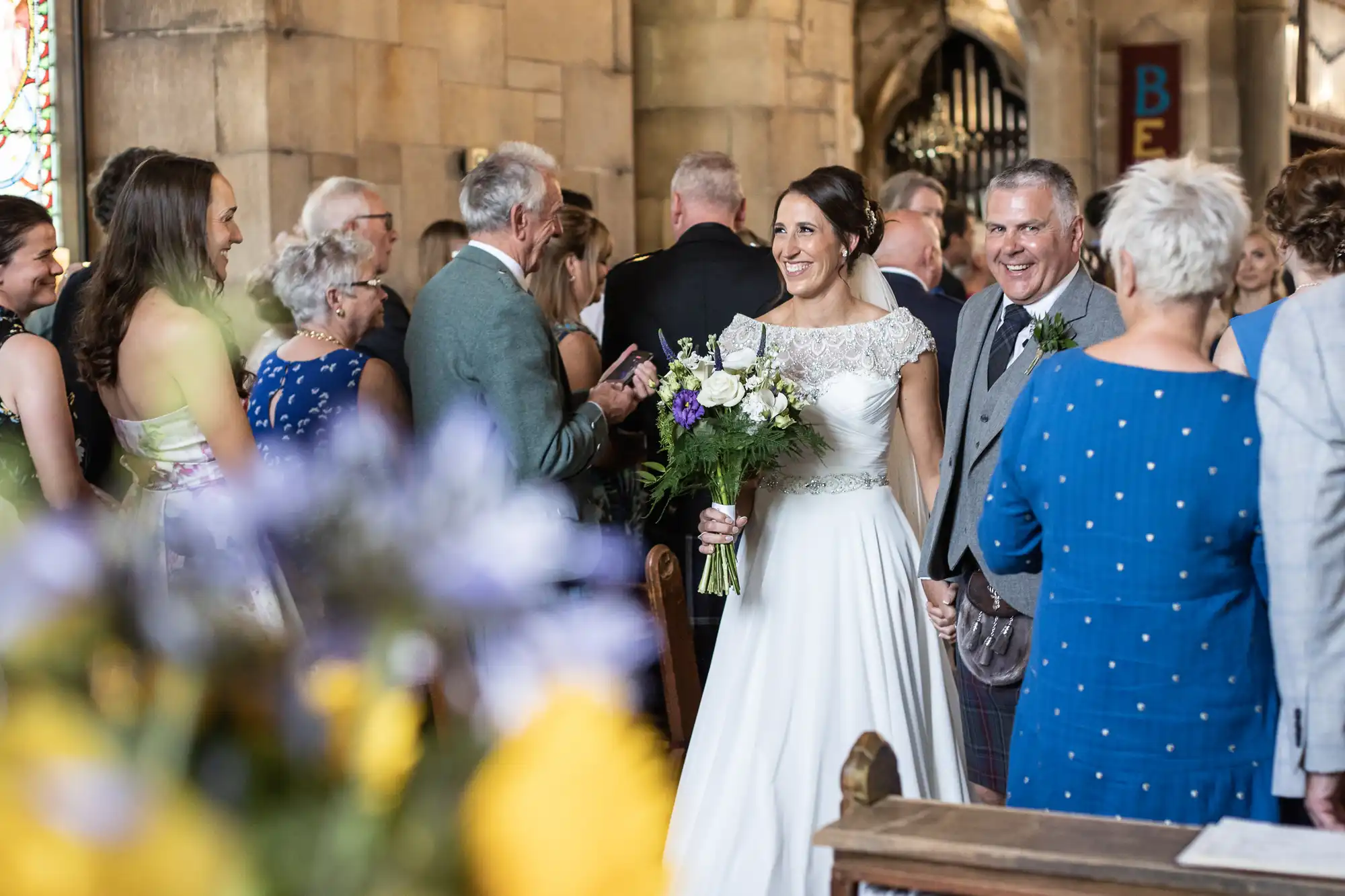 A bride and groom smile and hold hands while walking down the aisle in a church, surrounded by guests who are clapping and congratulating them. The bride holds a bouquet of flowers.