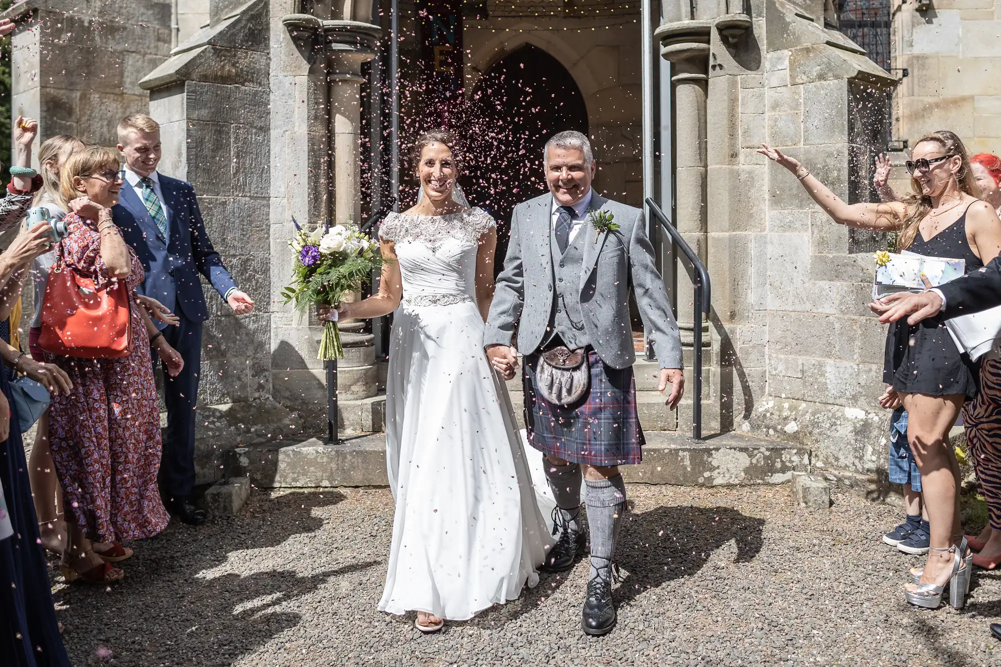 A bride and groom walk hand in hand outside a church, showered with confetti by guests. The groom is wearing a kilt and the bride is in a white wedding dress. Guests cheer and take photos.