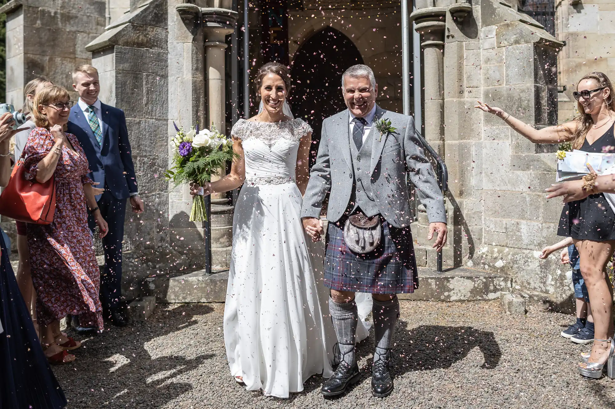 A bride in a white dress and groom in a kilt exit a stone building as guests throw confetti.