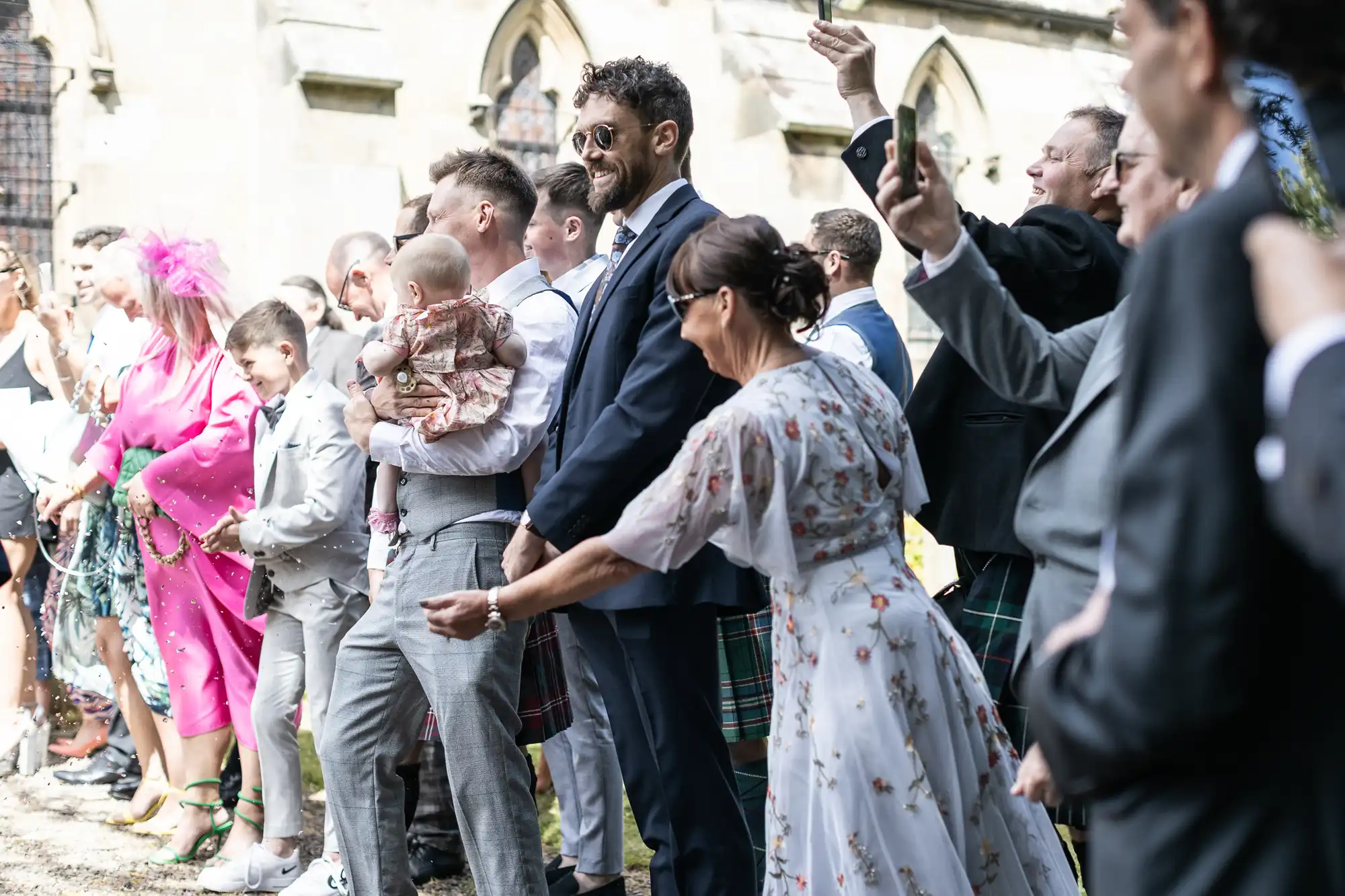 A group of people, including men in suits and some in kilts, and women in dresses, stand outside a building with a baby, taking photos and celebrating.