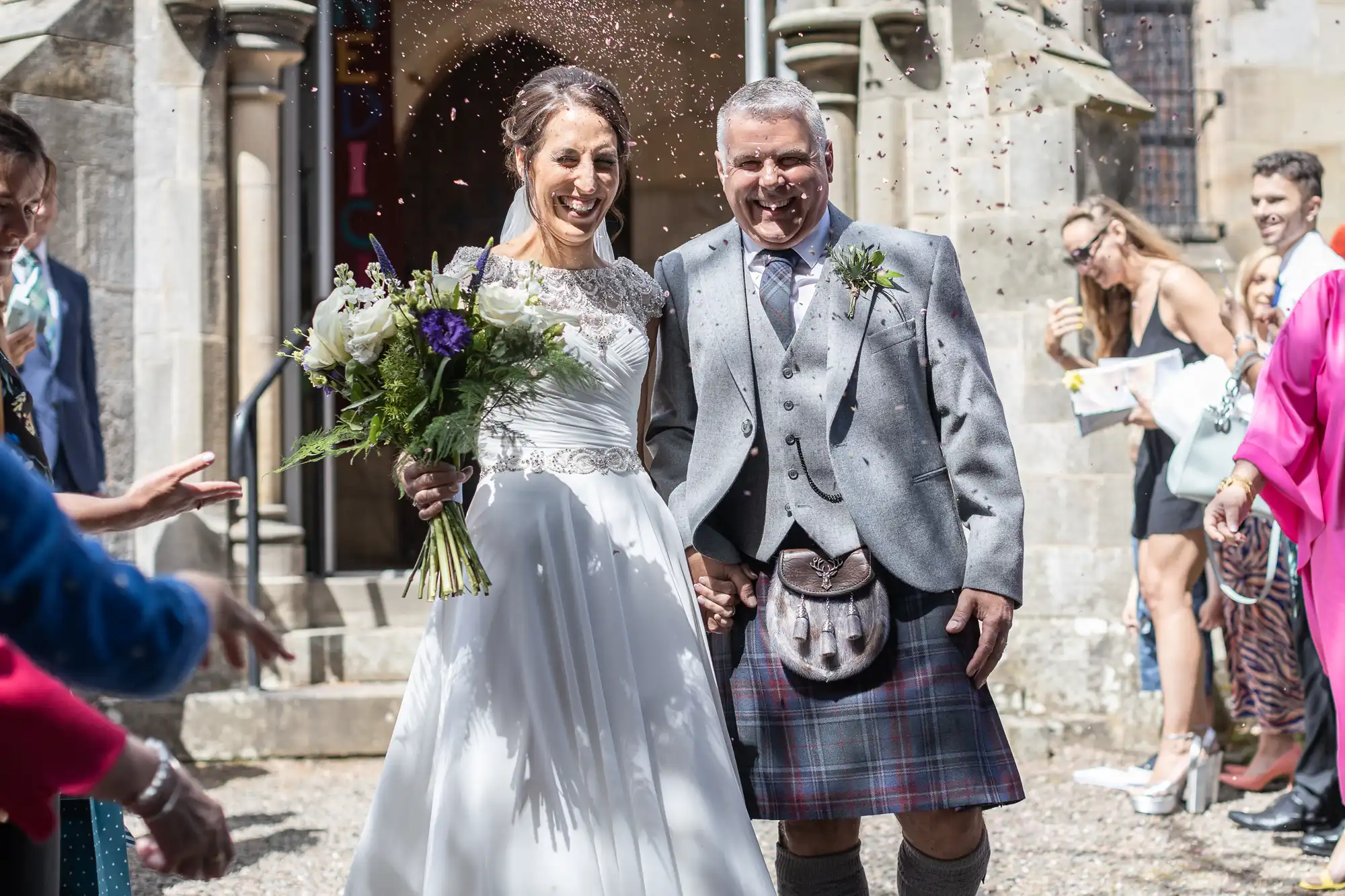 A bride in a white gown and groom in a kilt smile as they exit a building, holding a bouquet. Confetti is being thrown around them, and people are celebrating in the background.