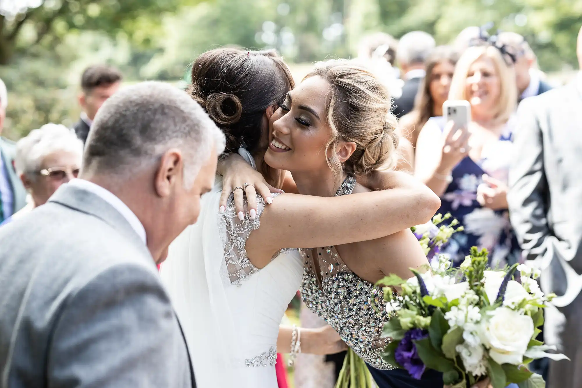 Two women embrace at an outdoor event. One holds a bouquet, and people in formal attire are visible in the background.