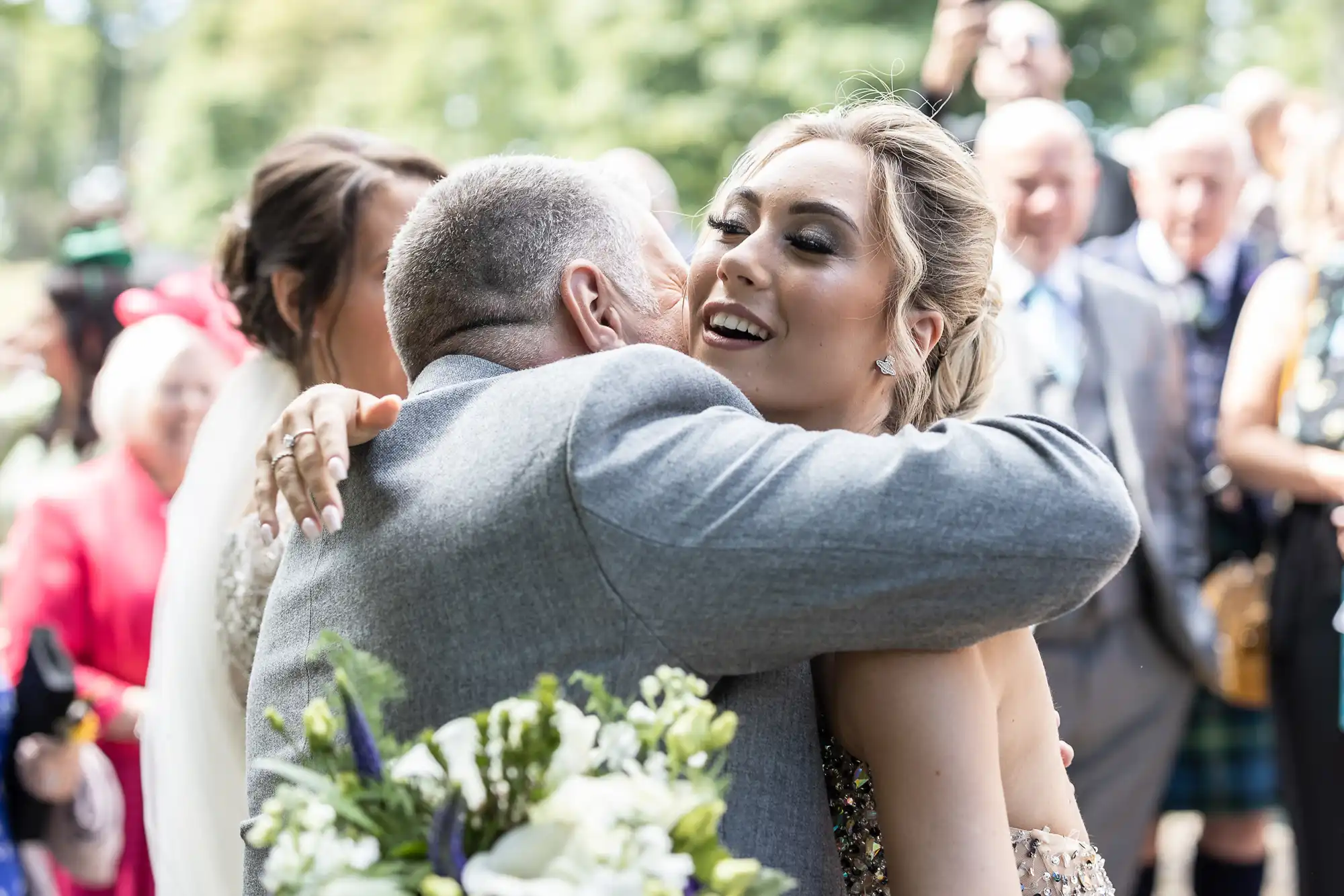 A man in a gray suit embraces a woman in a beaded dress, both smiling, during an outdoor event with people in the background.