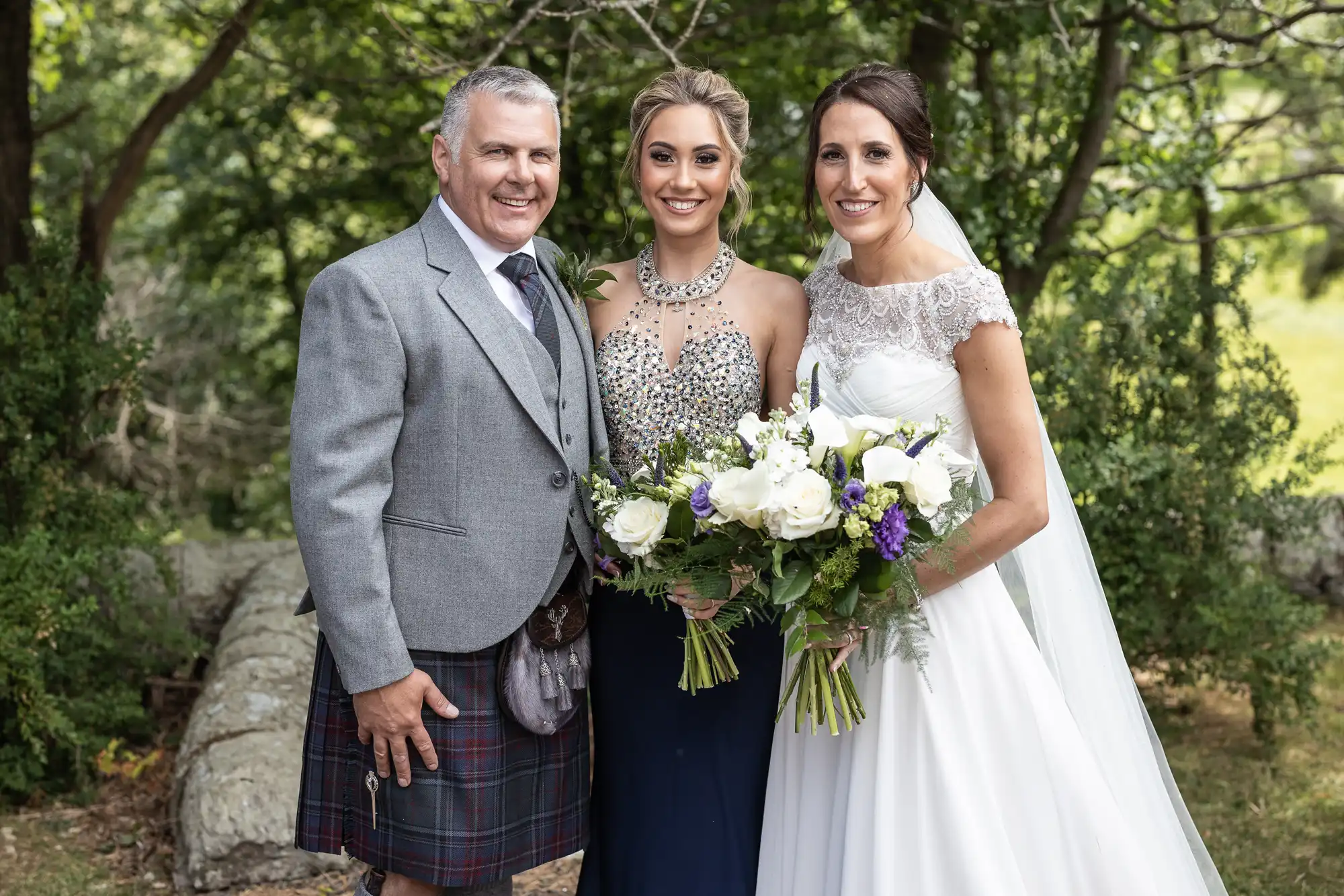 A man in a kilt, a woman in a beaded dress, and a woman in a bridal gown are standing outdoors, smiling, and holding bouquets.