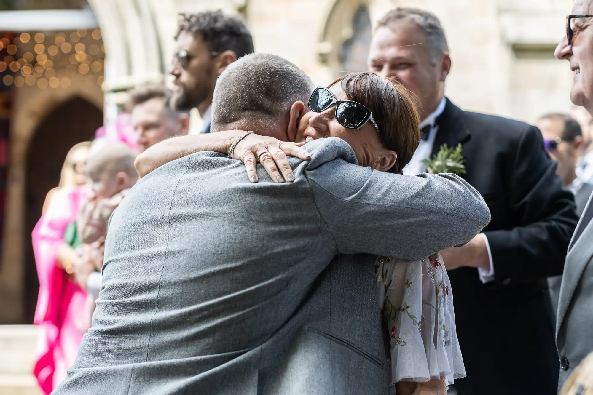 A man in a grey suit and a woman in sunglasses embrace at an outdoor event, with other people in formal attire standing in the background.