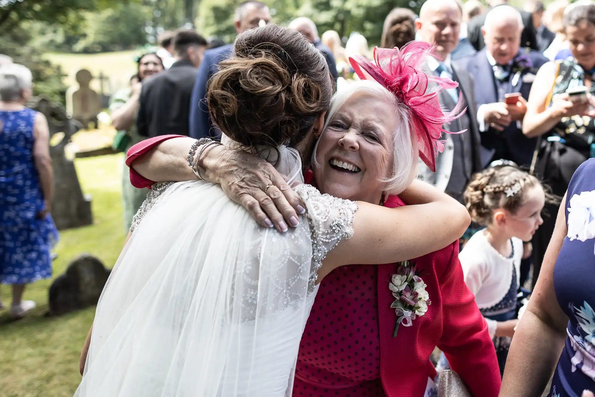 A bride with a lace veil hugs an older woman in a bright pink outfit and hat, surrounded by guests outdoors at a daytime event.