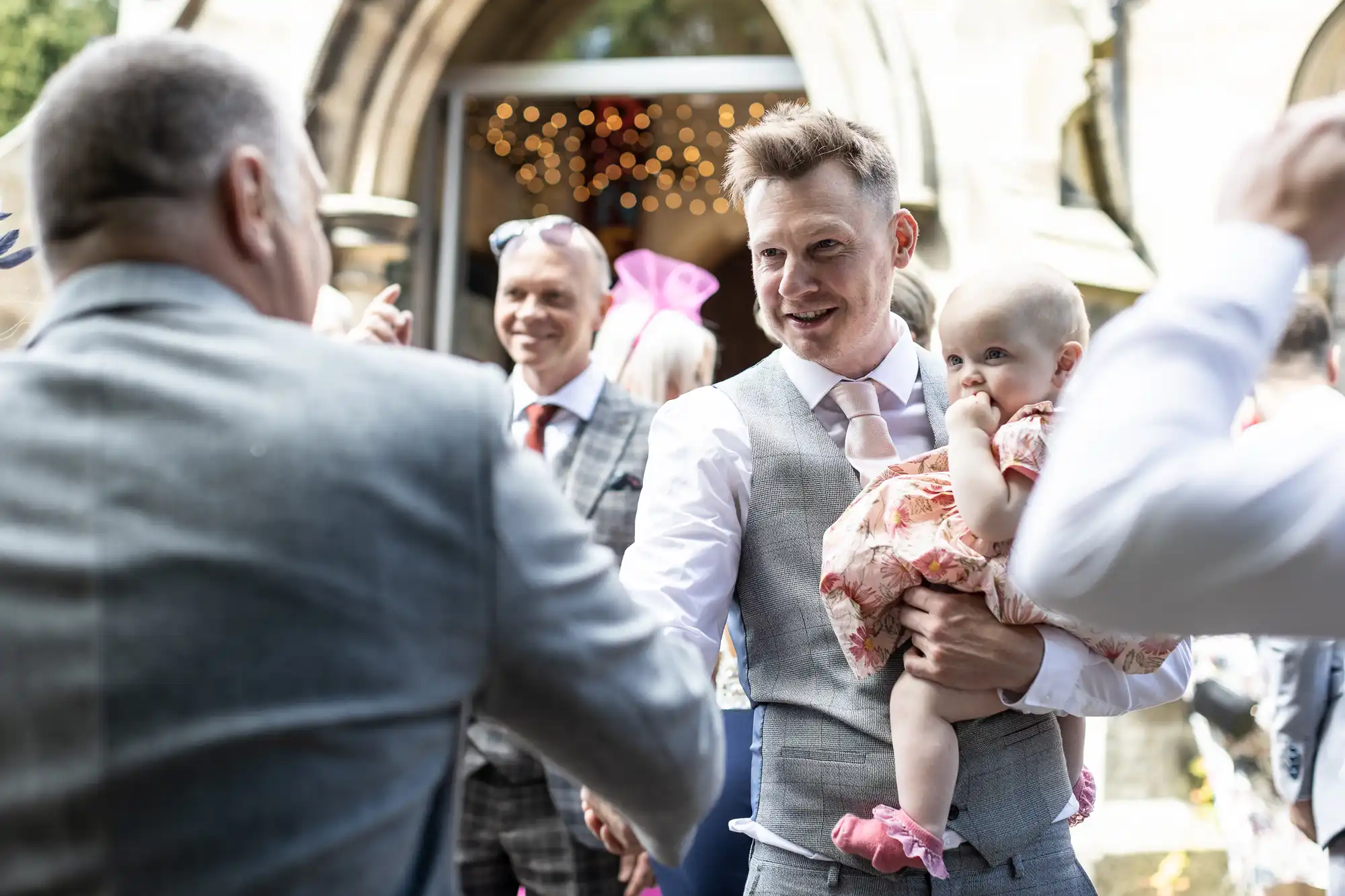 A man holding a baby shakes hands with another man outside a building, with other people in the background.