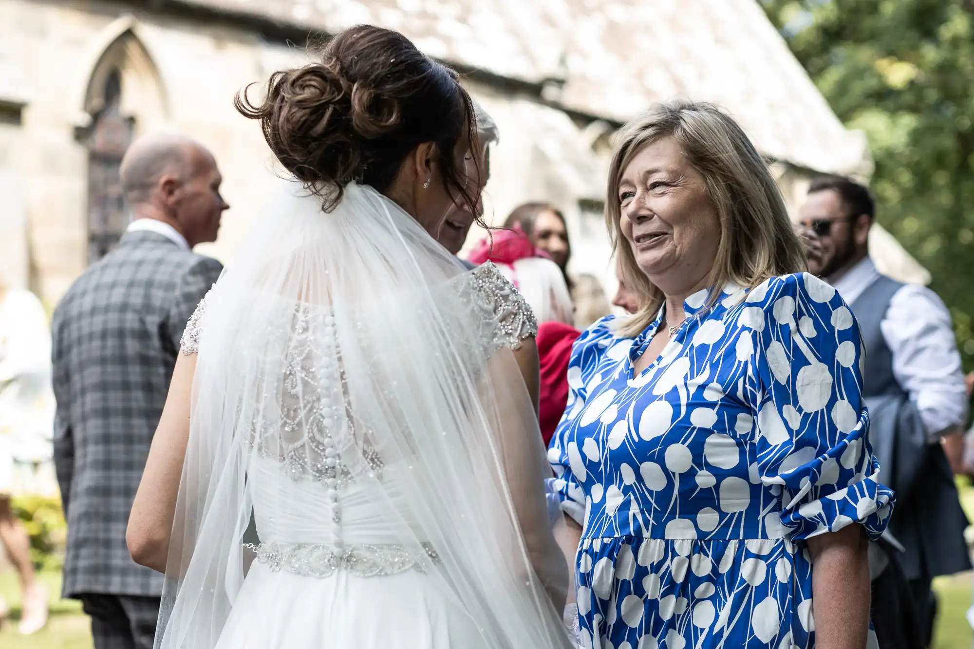 A bride in a white dress and veil speaks with a woman in a blue and white dress outdoors at a gathering. Other attendees are visible in the background.