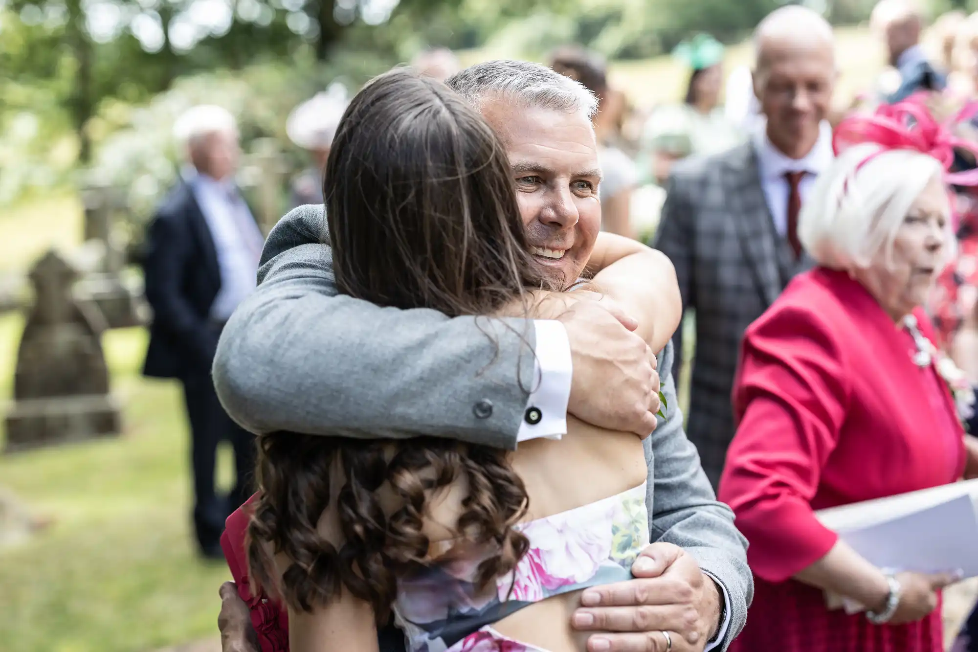 A man in a gray suit hugs a woman with long brown hair at an outdoor event. Several people in vibrant outfits are in the background.