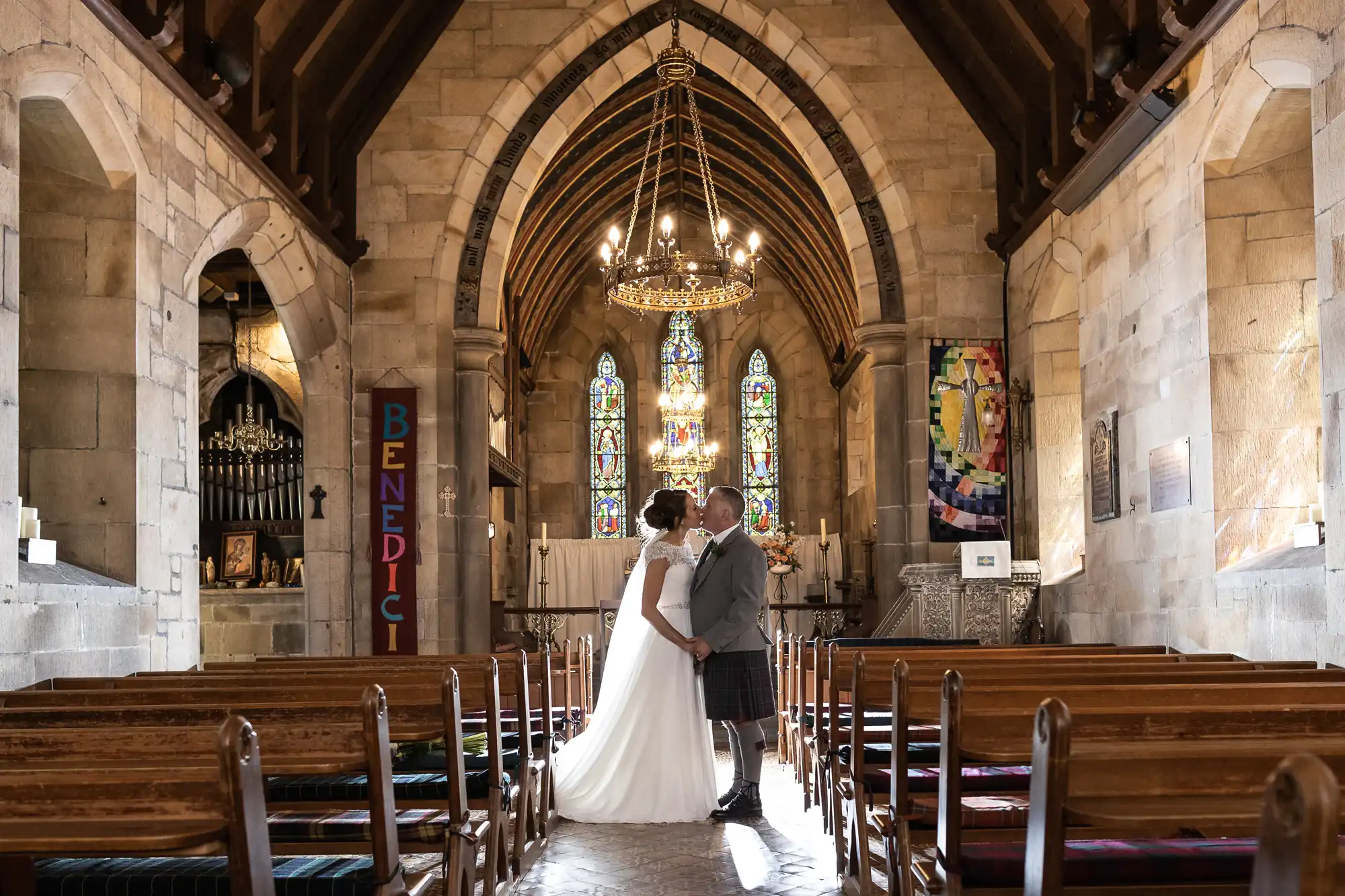 A couple, dressed in wedding attire, shares a kiss in the aisle of a stone church with an arched ceiling, ornate stained glass windows, and wooden pews.