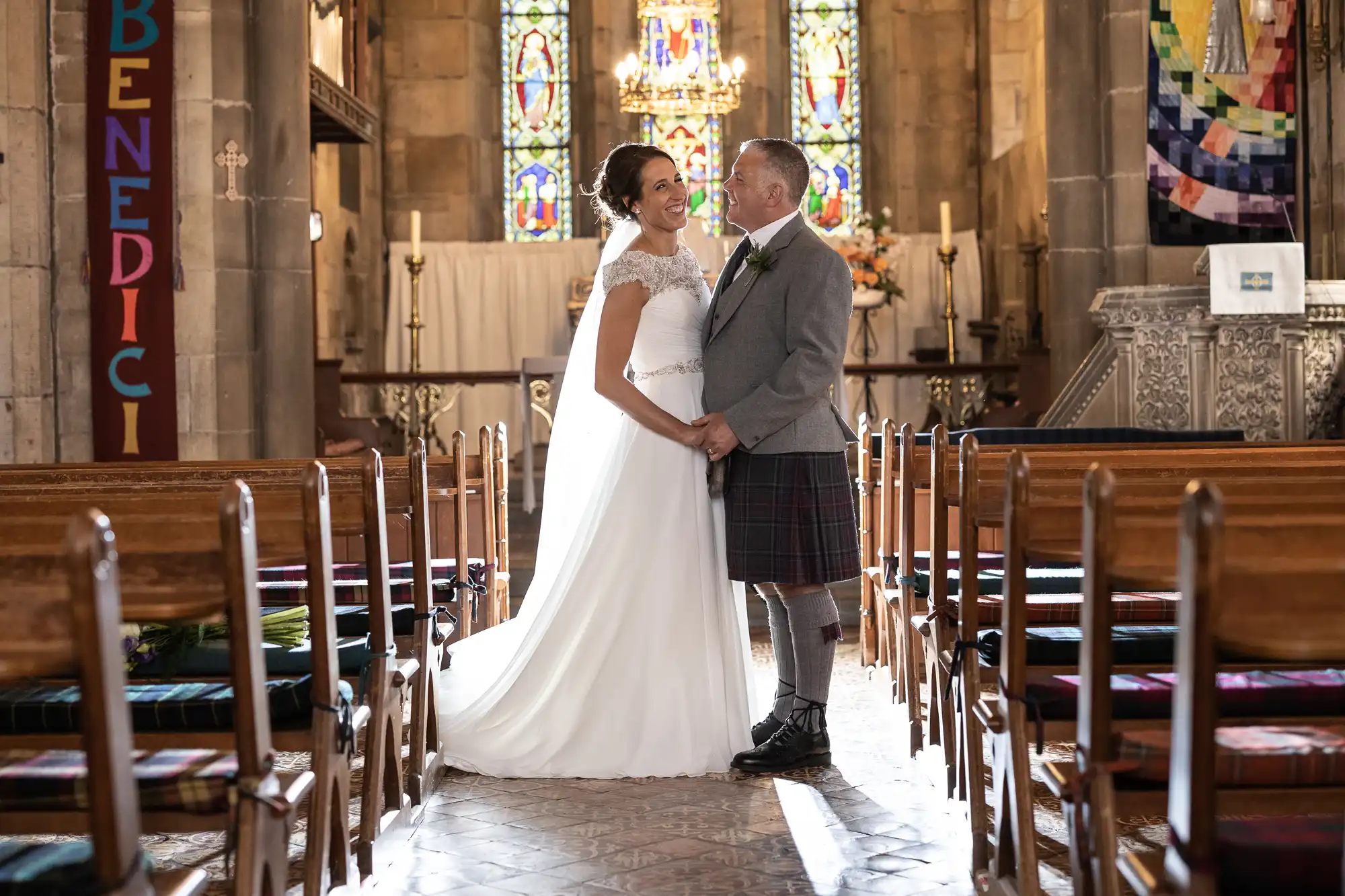 A couple dressed in wedding attire stand holding hands in the aisle of a church filled with wooden pews and stained glass windows, smiling at each other.