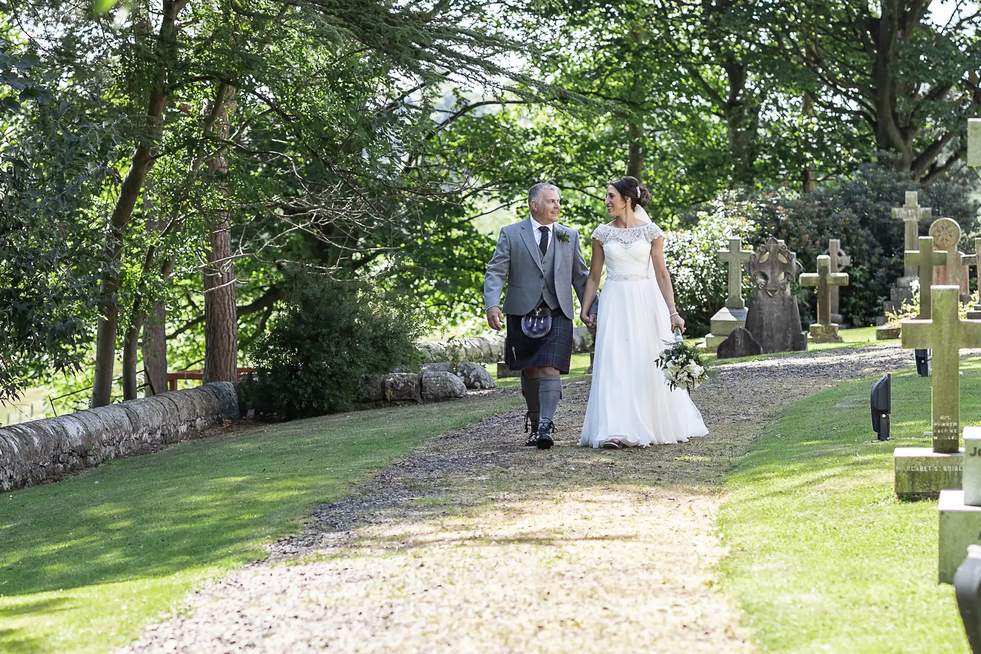 A bride in a white dress and a man in a kilt walk hand-in-hand along a garden path surrounded by greenery and gravestones.