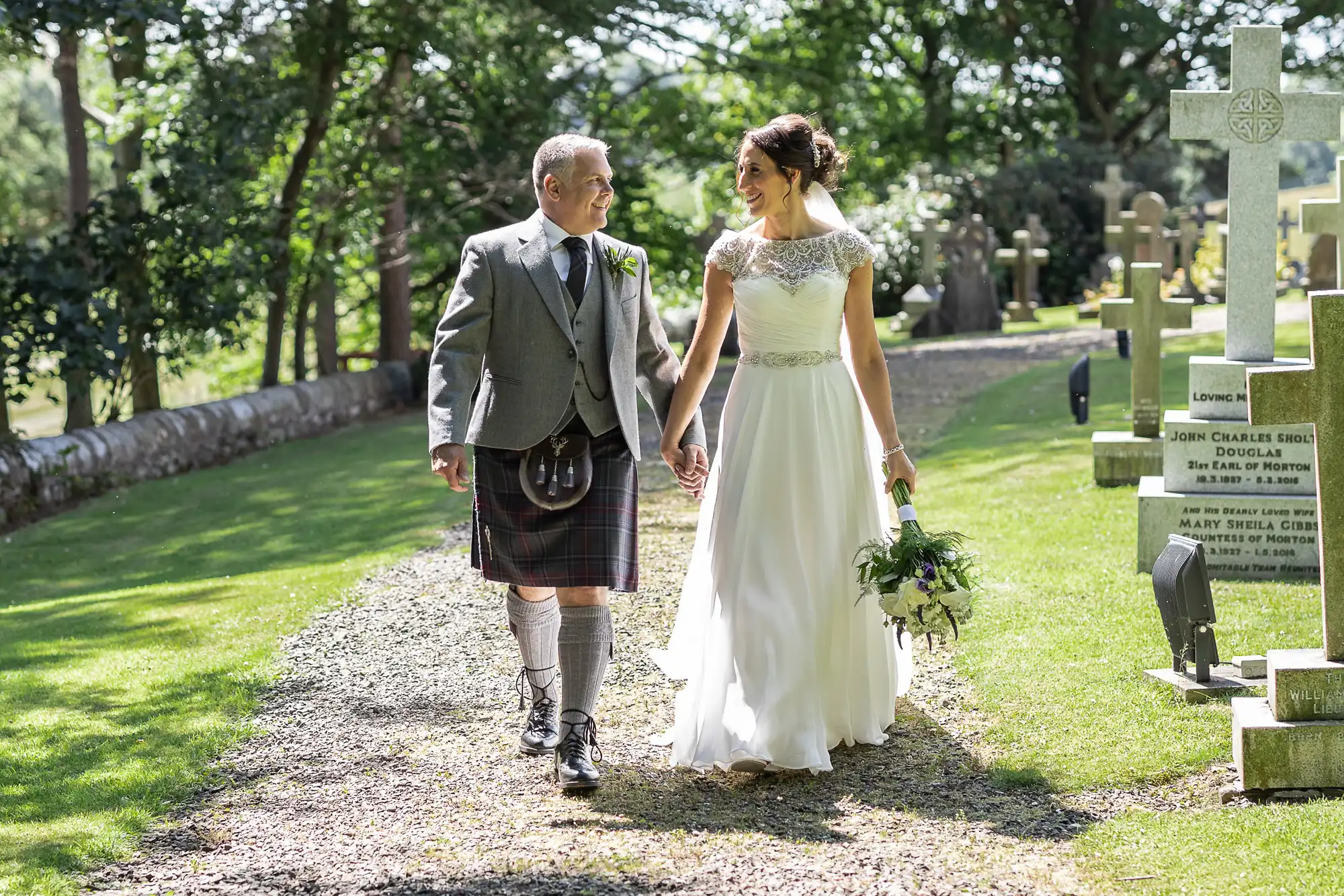 A bride and groom walk hand in hand along a gravel path in a cemetery surrounded by gravestones and greenery. The bride wears a white dress, and the groom wears a kilt with a grey jacket.