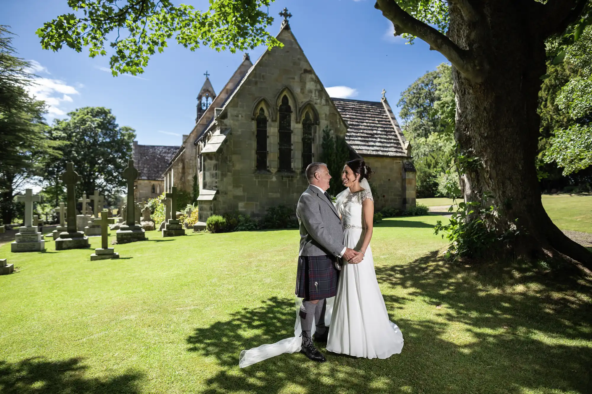 A couple in formal attire stand holding hands in front of a stone church and a large tree on a sunny day.