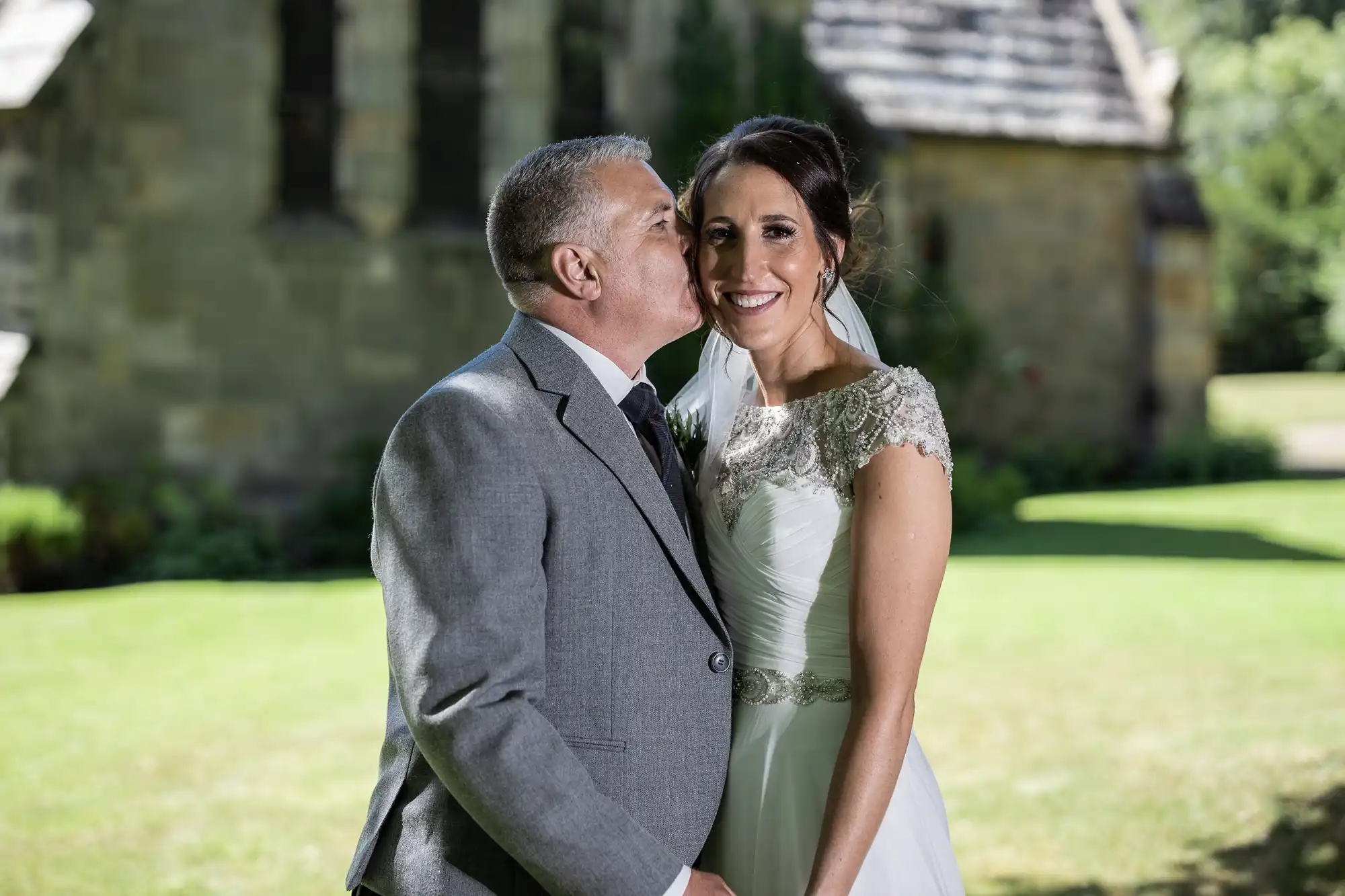 A man in a gray suit kisses a woman in a white wedding dress on the cheek. They stand outside in front of a stone building with green grass in the background.