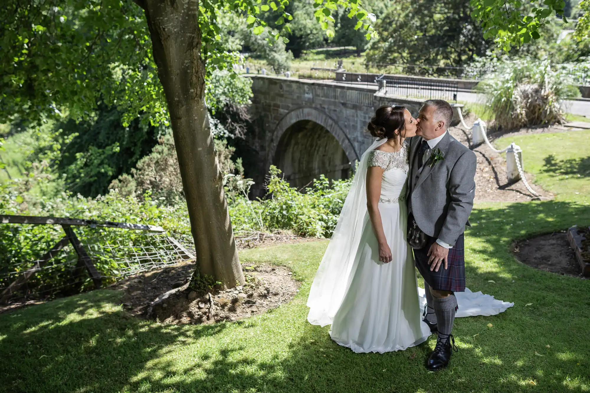 A bride and groom kiss under a tree near an old stone bridge, with greenery and a paved path in the background.