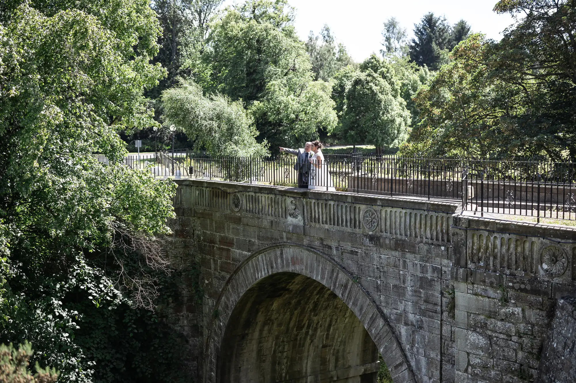 A couple in wedding attire stands on an old stone bridge surrounded by lush green trees.