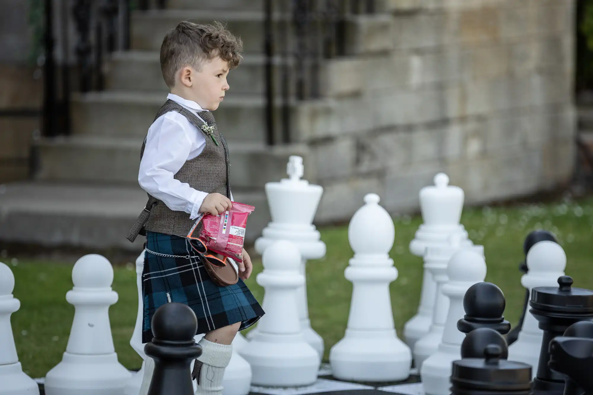 A young boy dressed in a kilt and vest holds a snack while walking on an oversized chessboard, surrounded by large chess pieces in a garden setting.