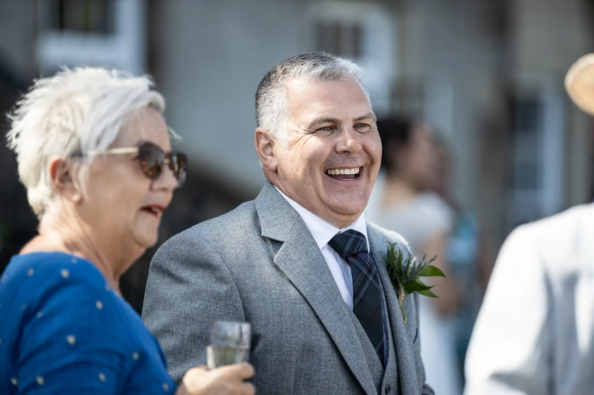 A man in a gray suit and tie stands outdoors, smiling broadly at an event. A woman with short white hair in a blue top and sunglasses is beside him, holding a glass.