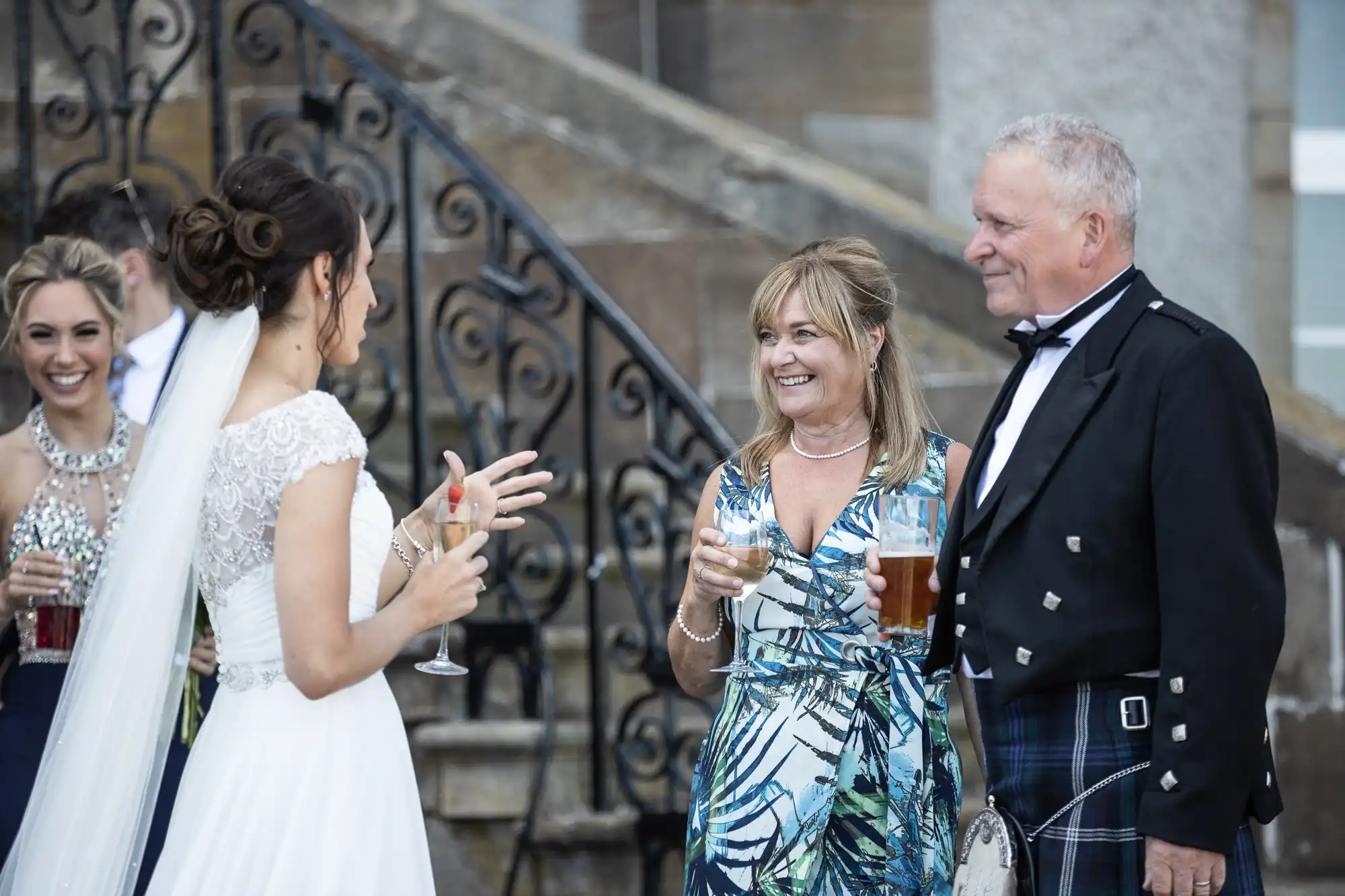 A bride in a white dress and veil converses with a man in a kilt and a woman in a floral dress; two women in formal attire smile in the background near a staircase with ornate railings.