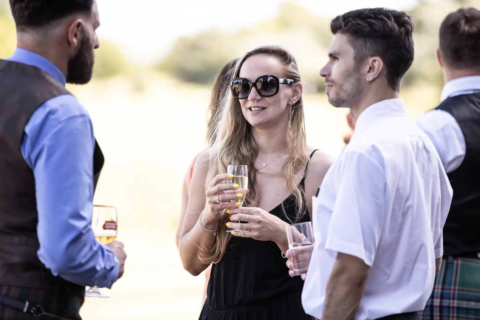 A group of people, including a woman holding a drink and wearing sunglasses, engages in conversation outdoors.