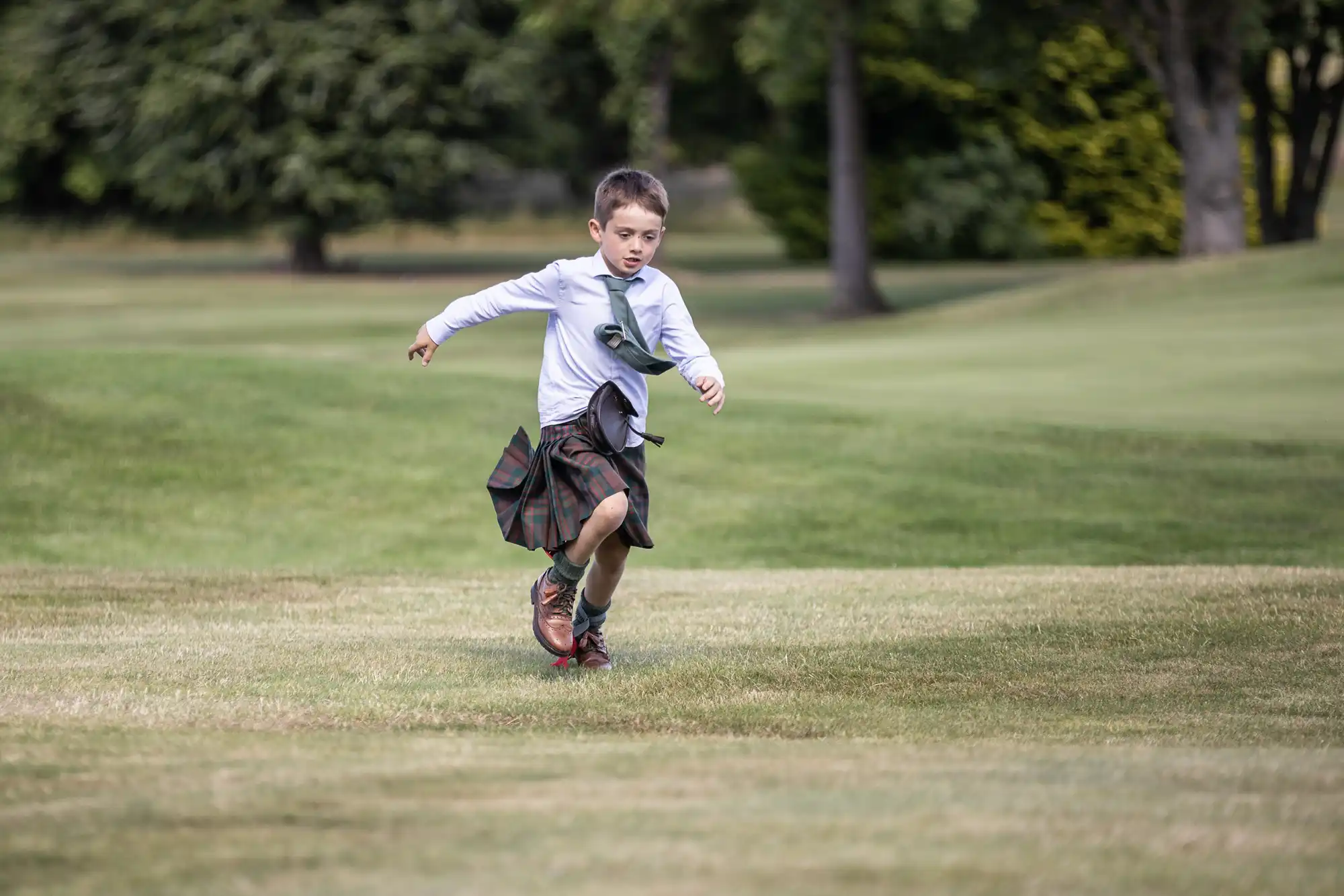 A young boy wearing a kilt and long-sleeved shirt runs across a grassy field with trees in the background.