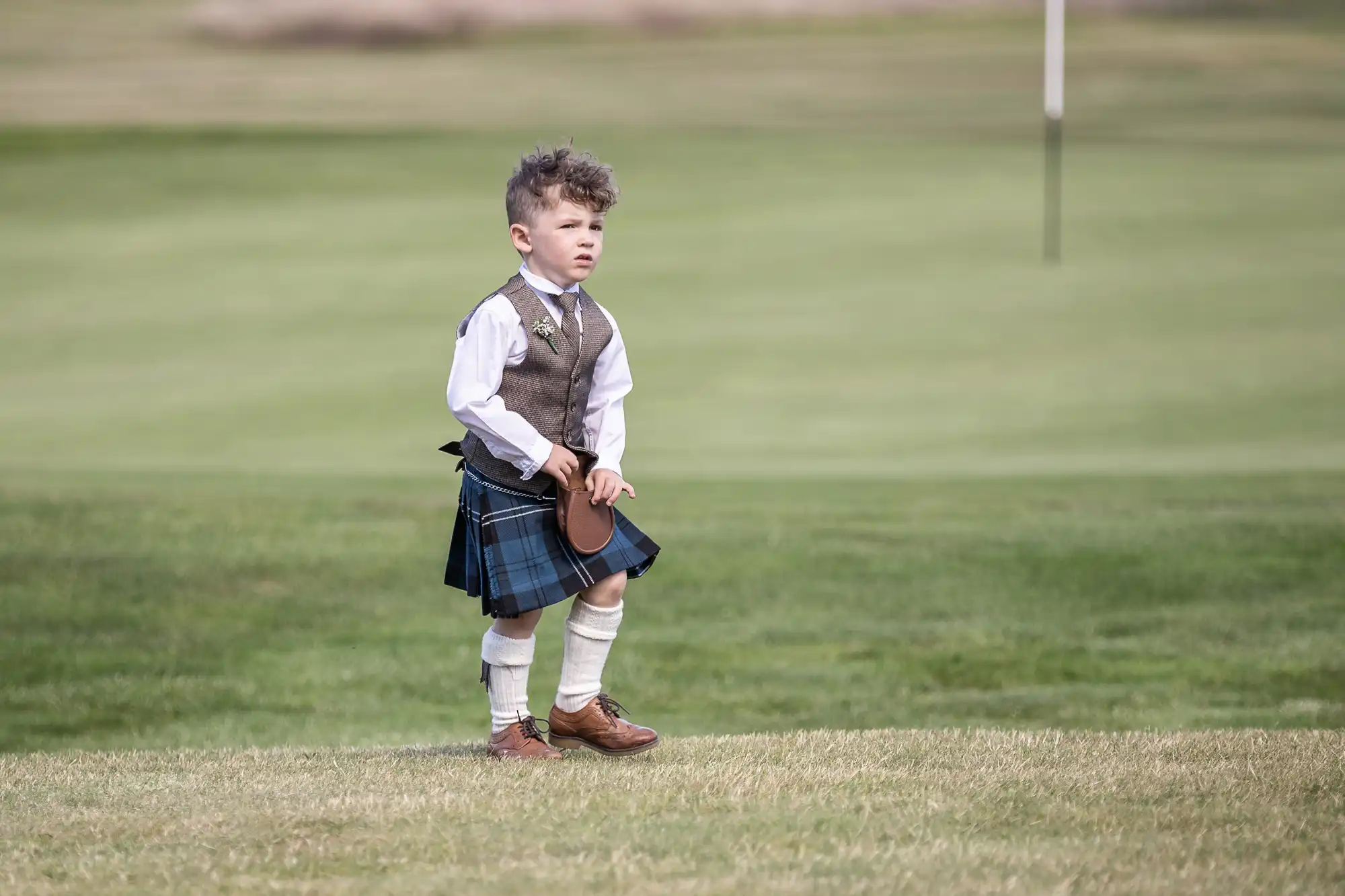 A young child stands on a grassy field, wearing a traditional plaid kilt, vest, white shirt, knee-high socks, and brown shoes, holding a small hat in one hand.