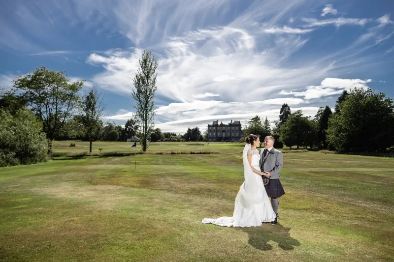 Wedding photographer at the Dalmahoy Hotel: A couple dressed in wedding attire stands on a grassy field in front of a large building. The bride is in a white dress, and the groom wears a kilt. The sky is filled with scattered white clouds.