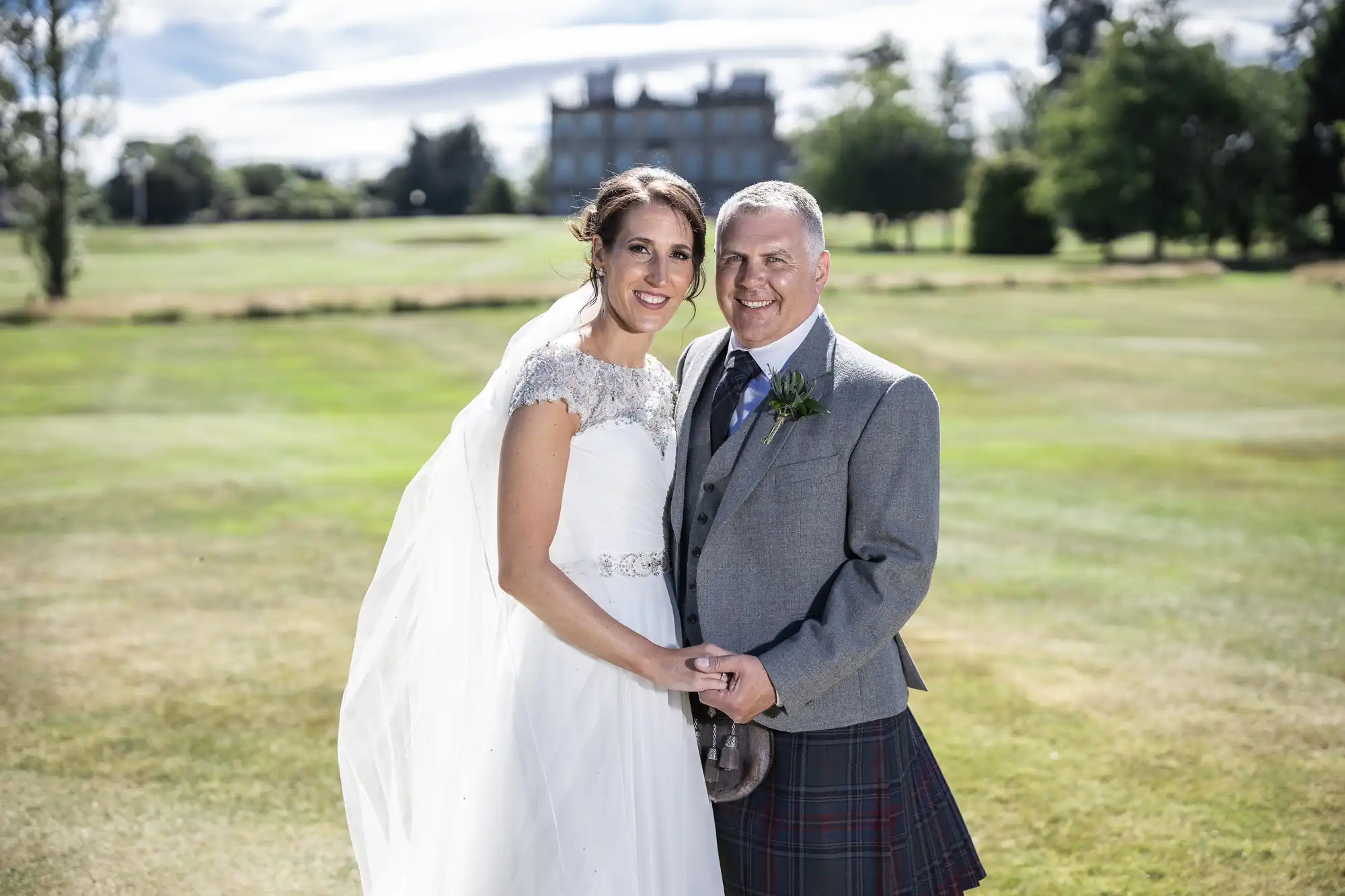 A couple in wedding attire stands outdoors, smiling. The bride wears a white dress and veil, while the groom wears a grey suit with a tartan kilt. A large building and trees are visible in the background.