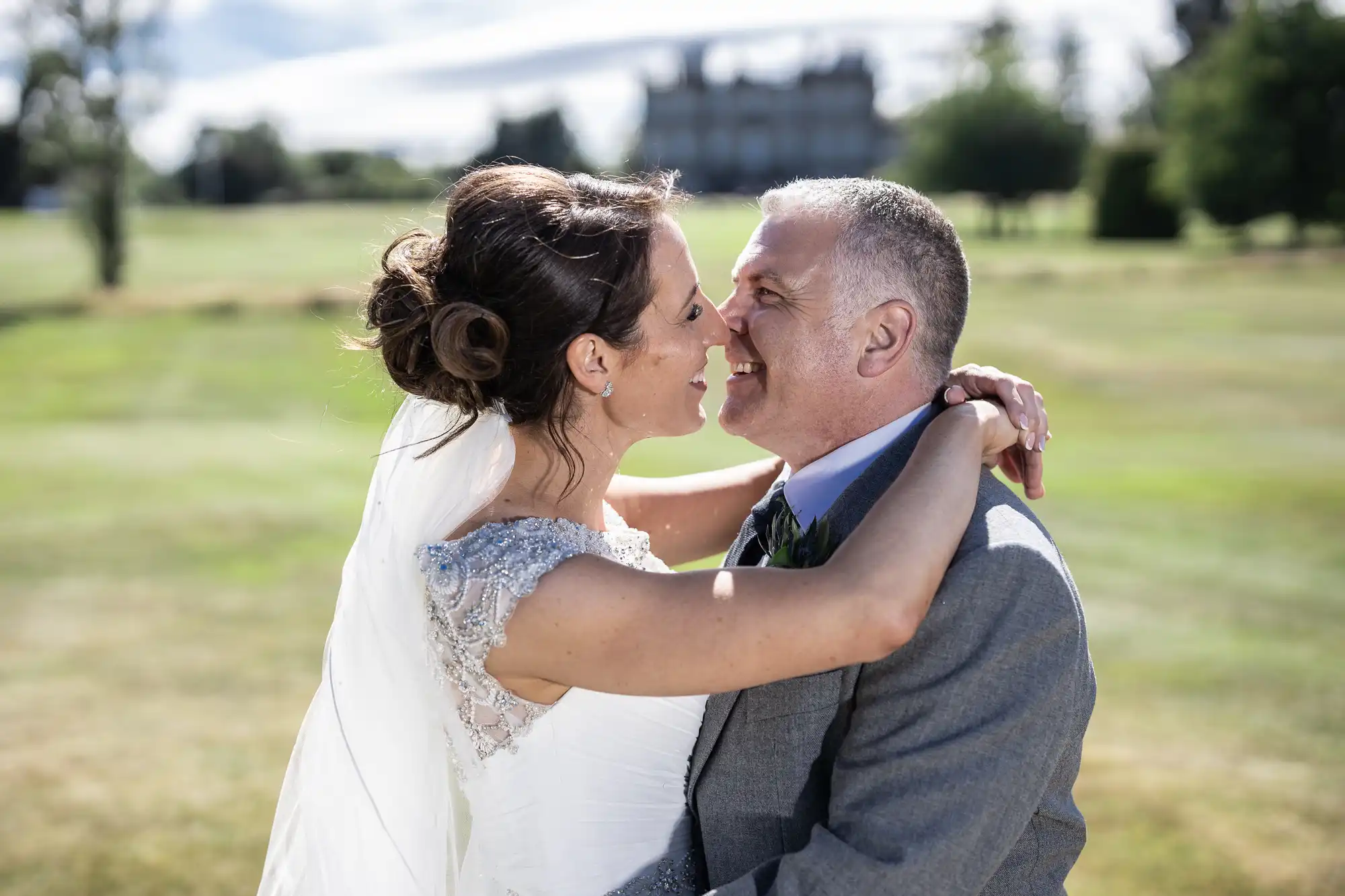 A bride and groom embrace and smile at each other outdoors, with a vast green landscape and a large building in the background.