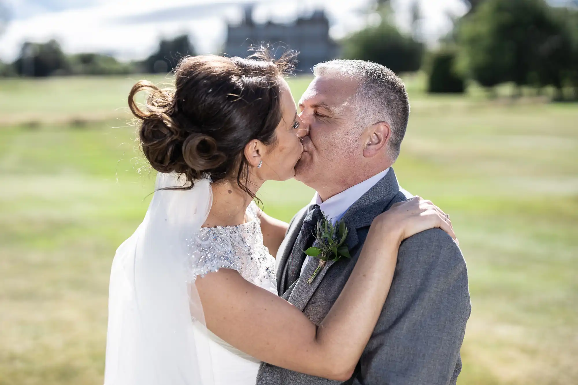 Bride and groom share a kiss outdoors on a sunny day, with an expansive green field and large building in the background.