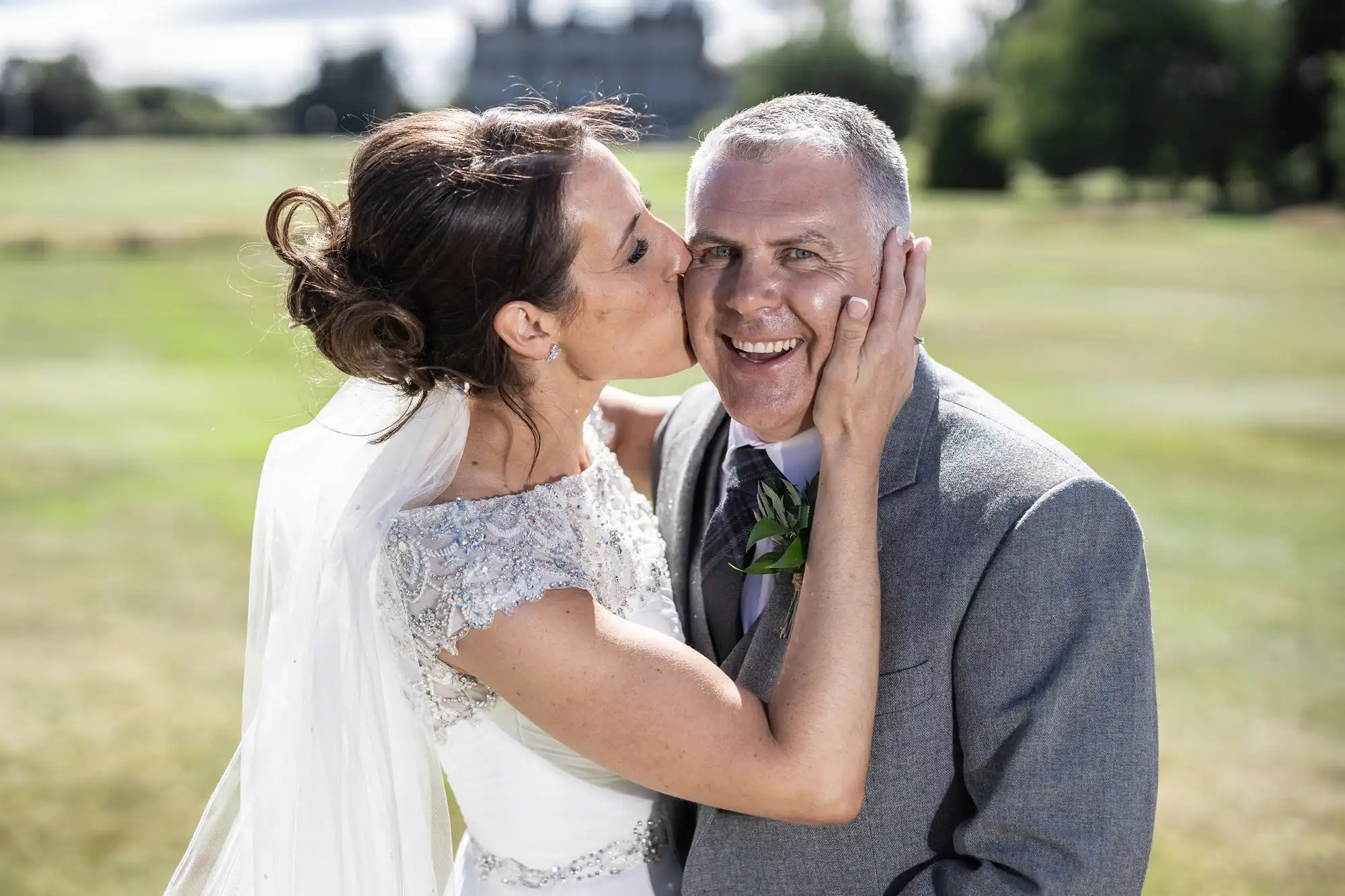 A bride in a white dress and veil kisses a smiling groom in a gray suit on the cheek in an outdoor setting.