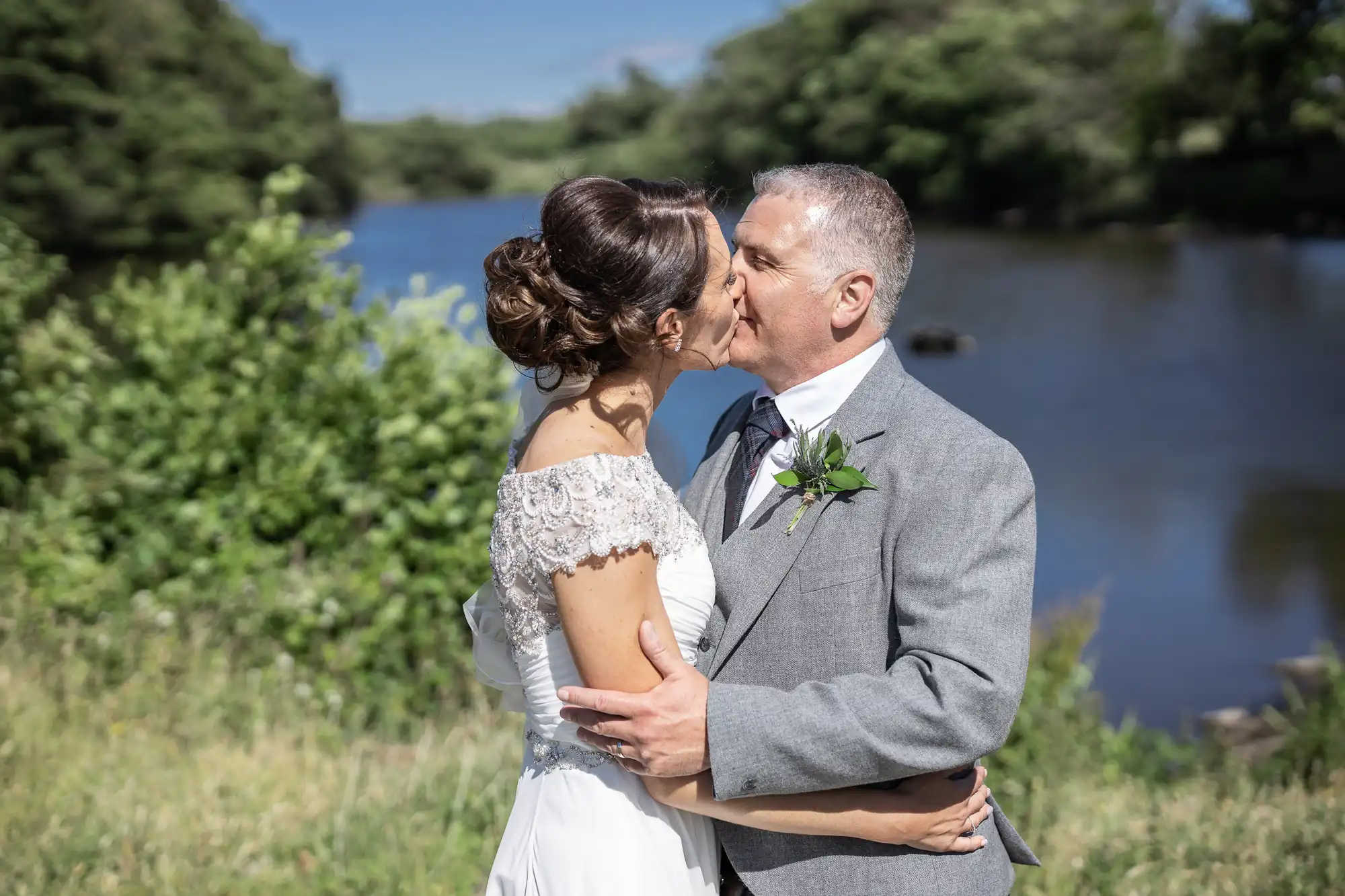 A couple in wedding attire kisses while standing outdoors near a river, with greenery and trees in the background.