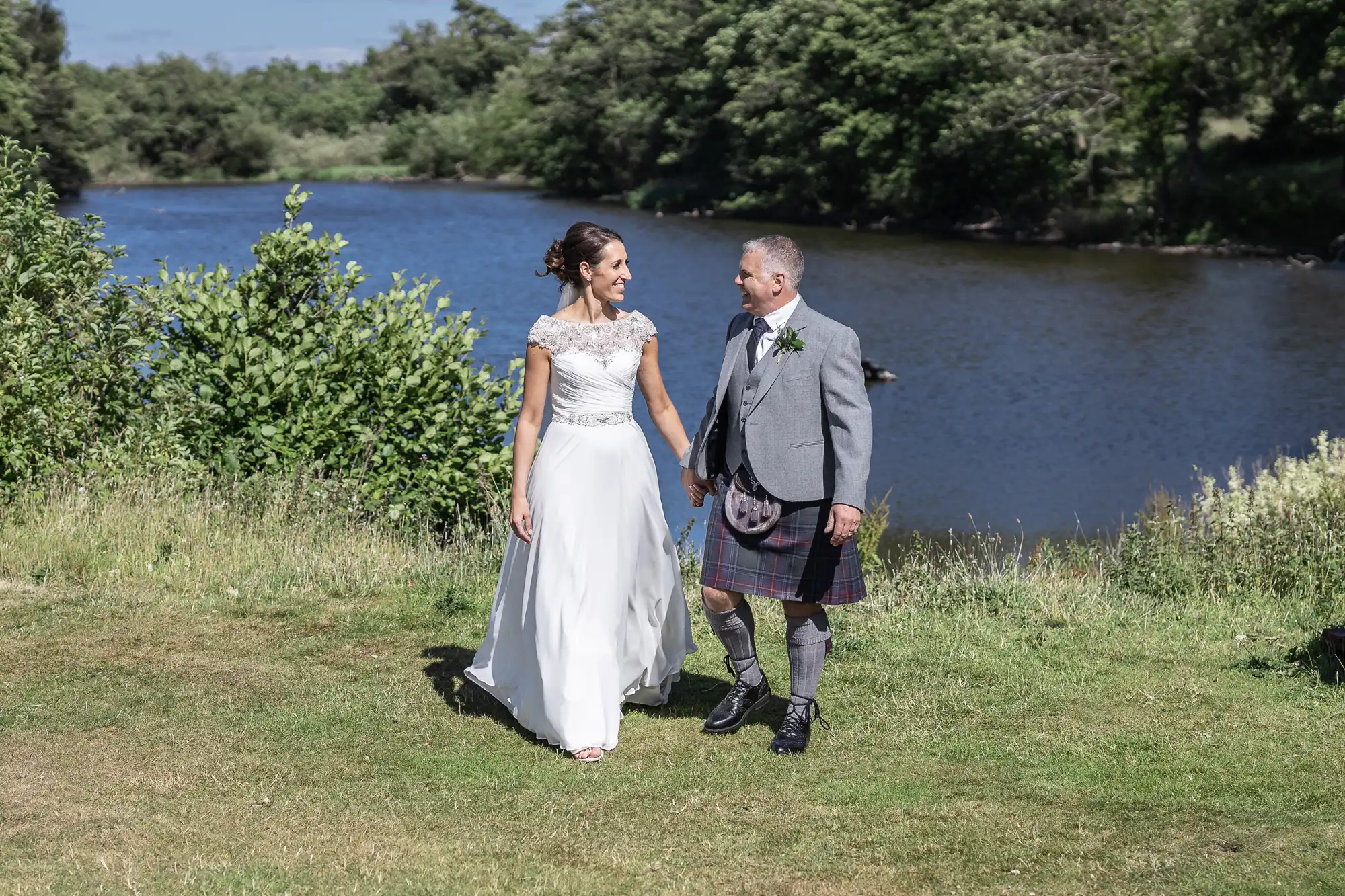 A bride and groom, with the groom in a kilt, walk hand in hand on a grassy area by a river on a sunny day.