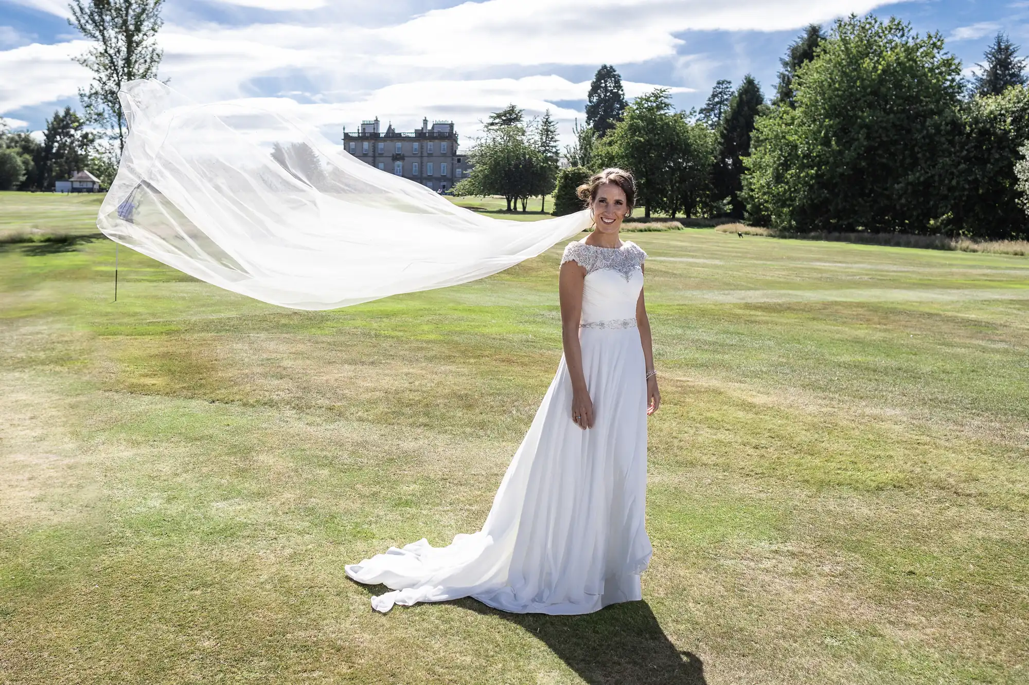 A bride in a white gown stands on a grassy field with trees and a building in the background, her veil flowing in the wind.