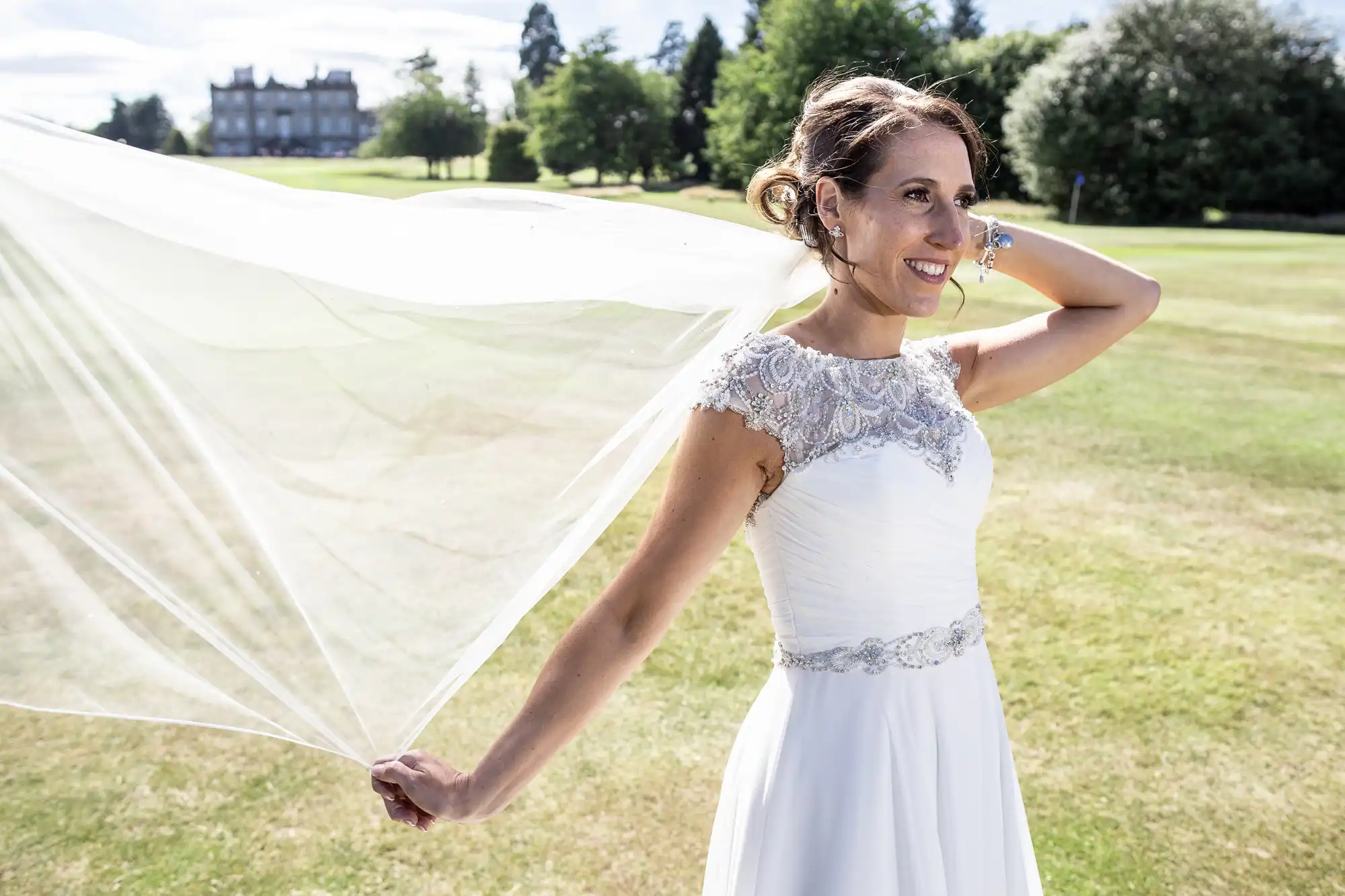 A smiling woman in a white wedding dress holds her veil while standing on a grassy field with trees and a large building in the background.