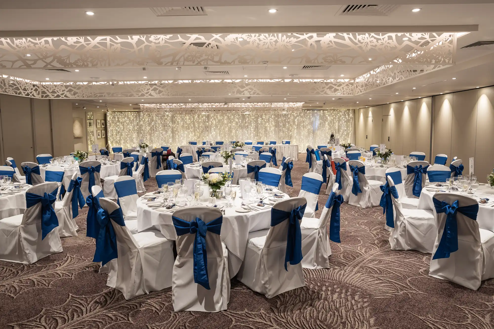 A decorated wedding reception hall with round tables set for dining, white chairs covered with blue ribbon sashes, floral centerpieces, and a backdrop of string lights.