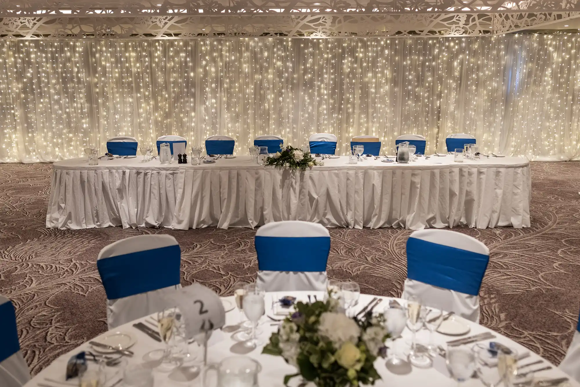 A long, elegantly decorated wedding table with white tablecloth and blue chair covers, set in front of a background with hanging string lights.