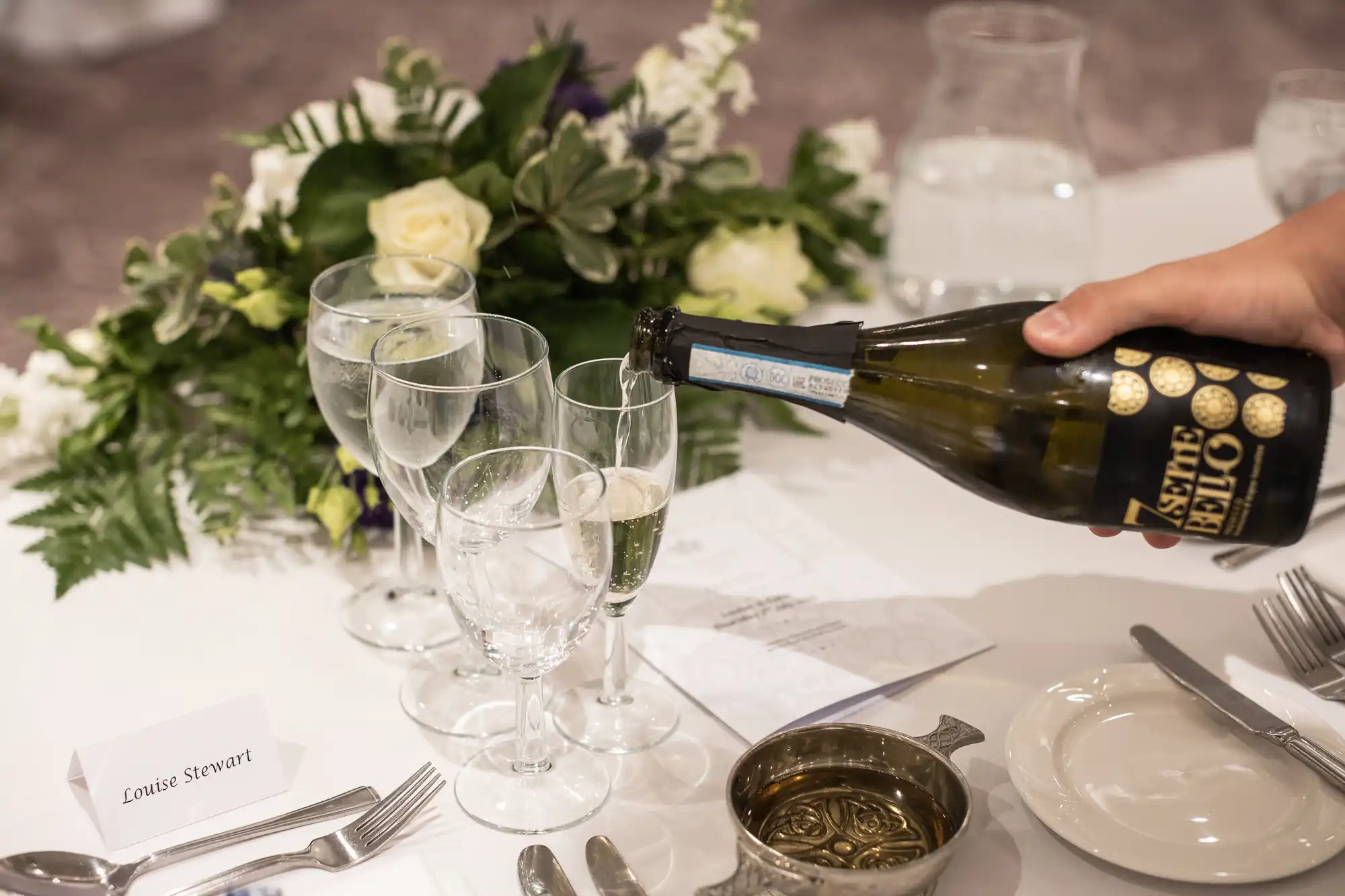 A person pours champagne into a glass on a table set with flowers, cutlery, plates, and a "Louise Stewart" name card, indicating a formal event or celebration.