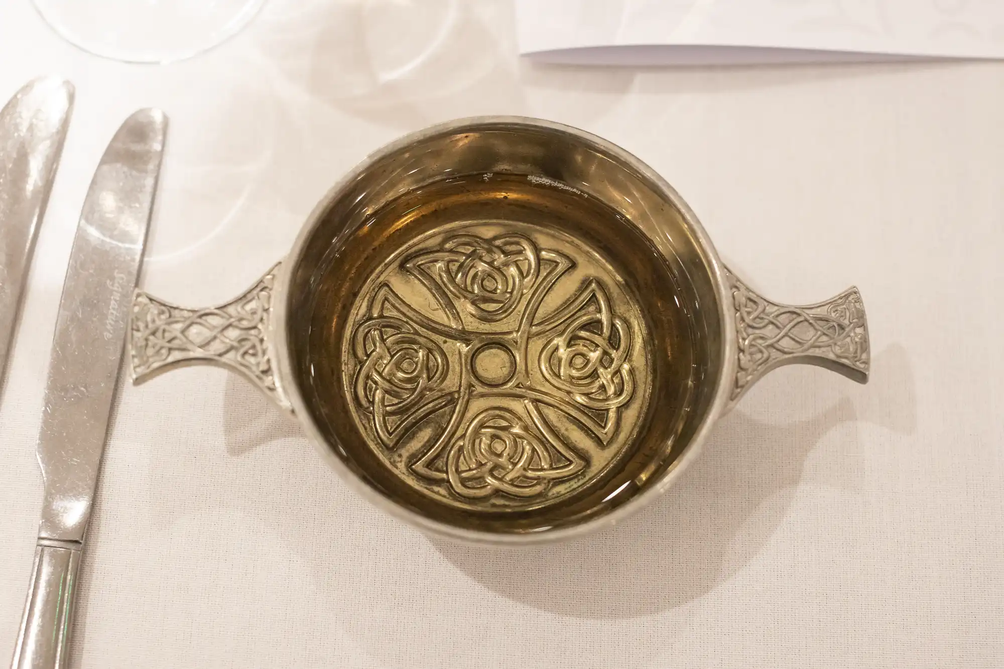 A small, intricate silver bowl with two handles, featuring a detailed rose pattern inside, placed on a white cloth next to a knife.