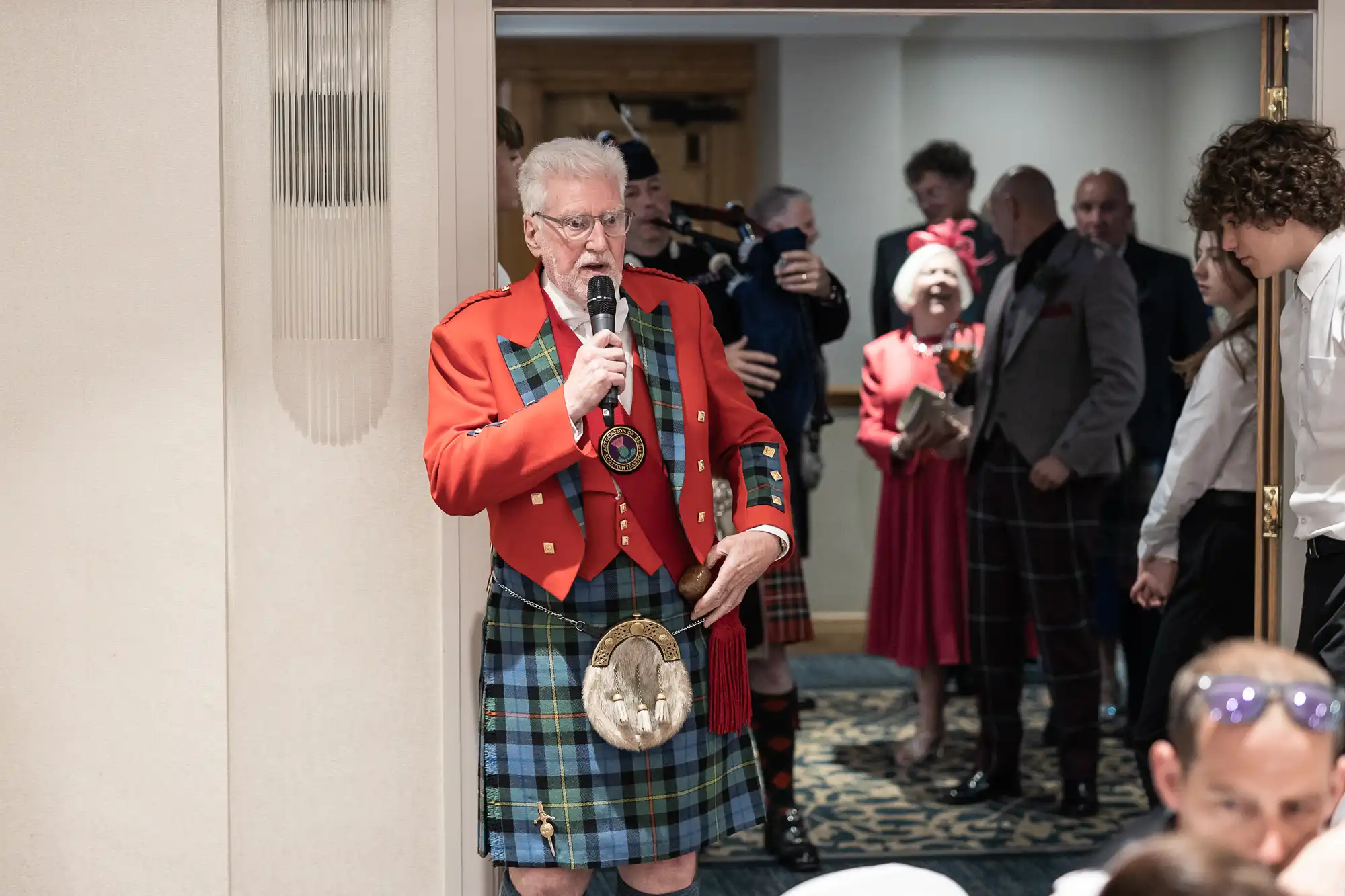 An elderly man in a red jacket and tartan kilt speaks into a microphone. People, including a woman in red, are in the background, appearing to attend an event or celebration.