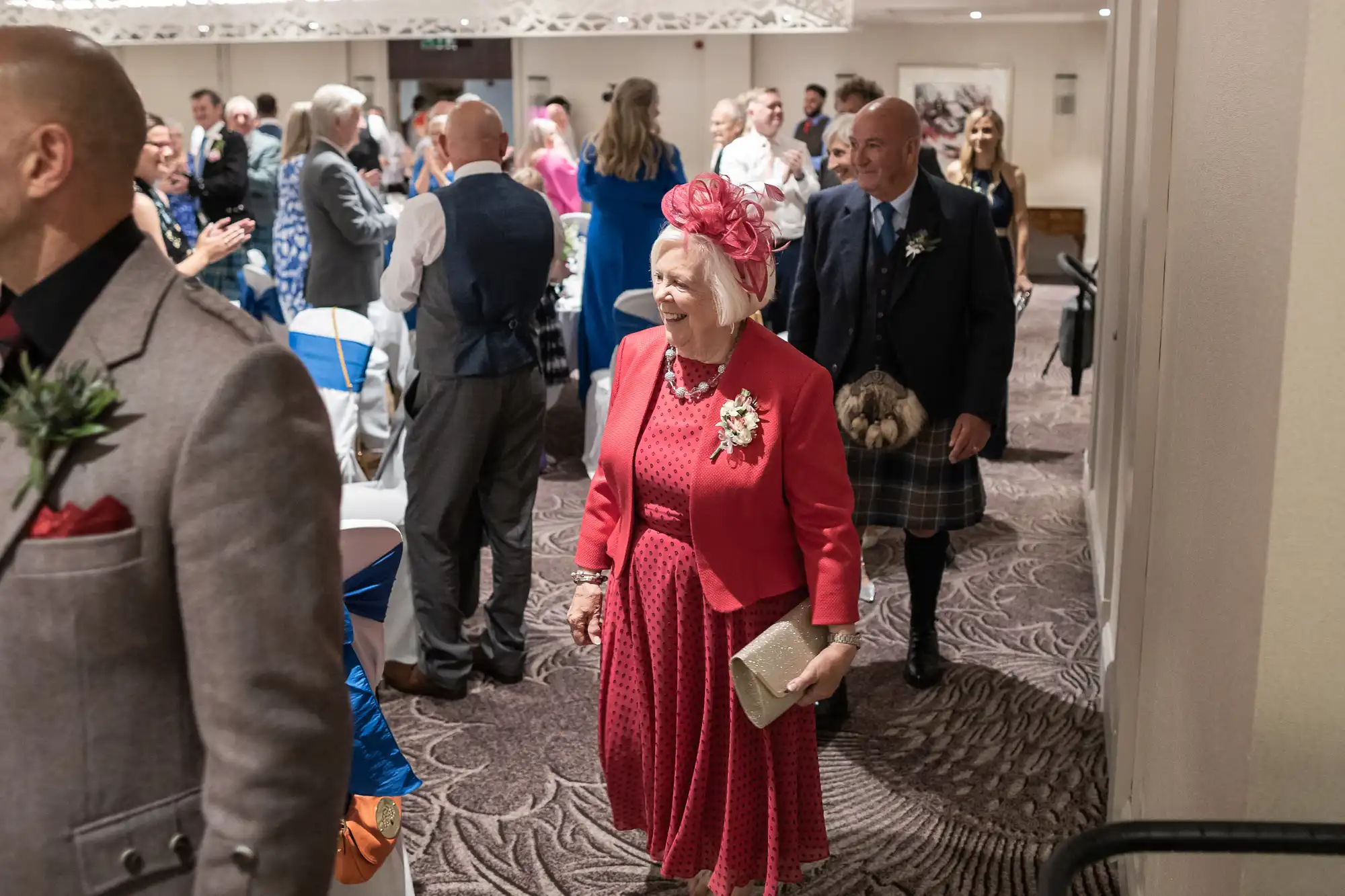 An older woman in a red outfit and matching fascinator smiles while walking through a room filled with people at a formal event.