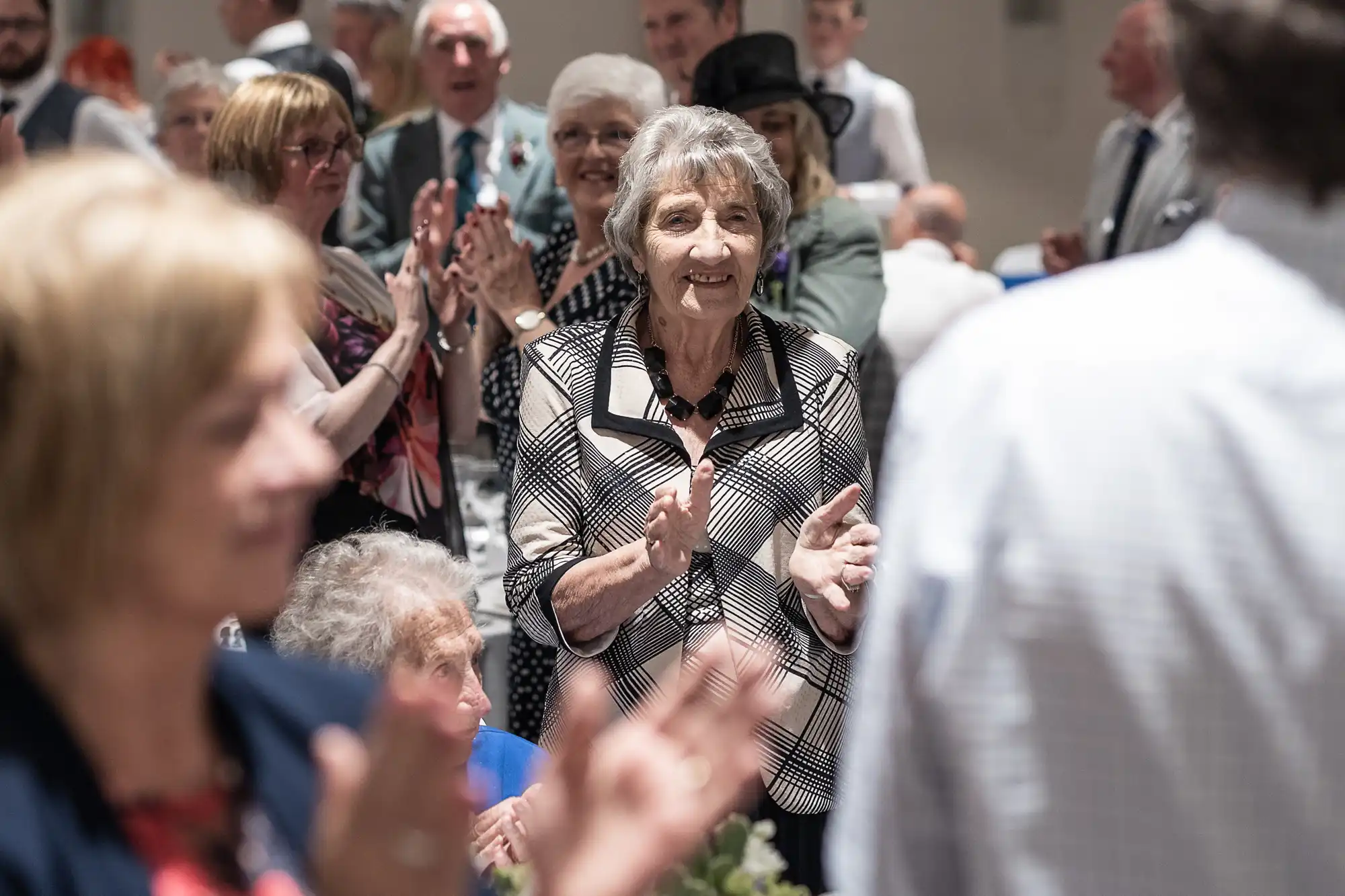 Elderly woman standing and clapping in a crowd of seated and standing people, who are also applauding in a well-lit gathering.